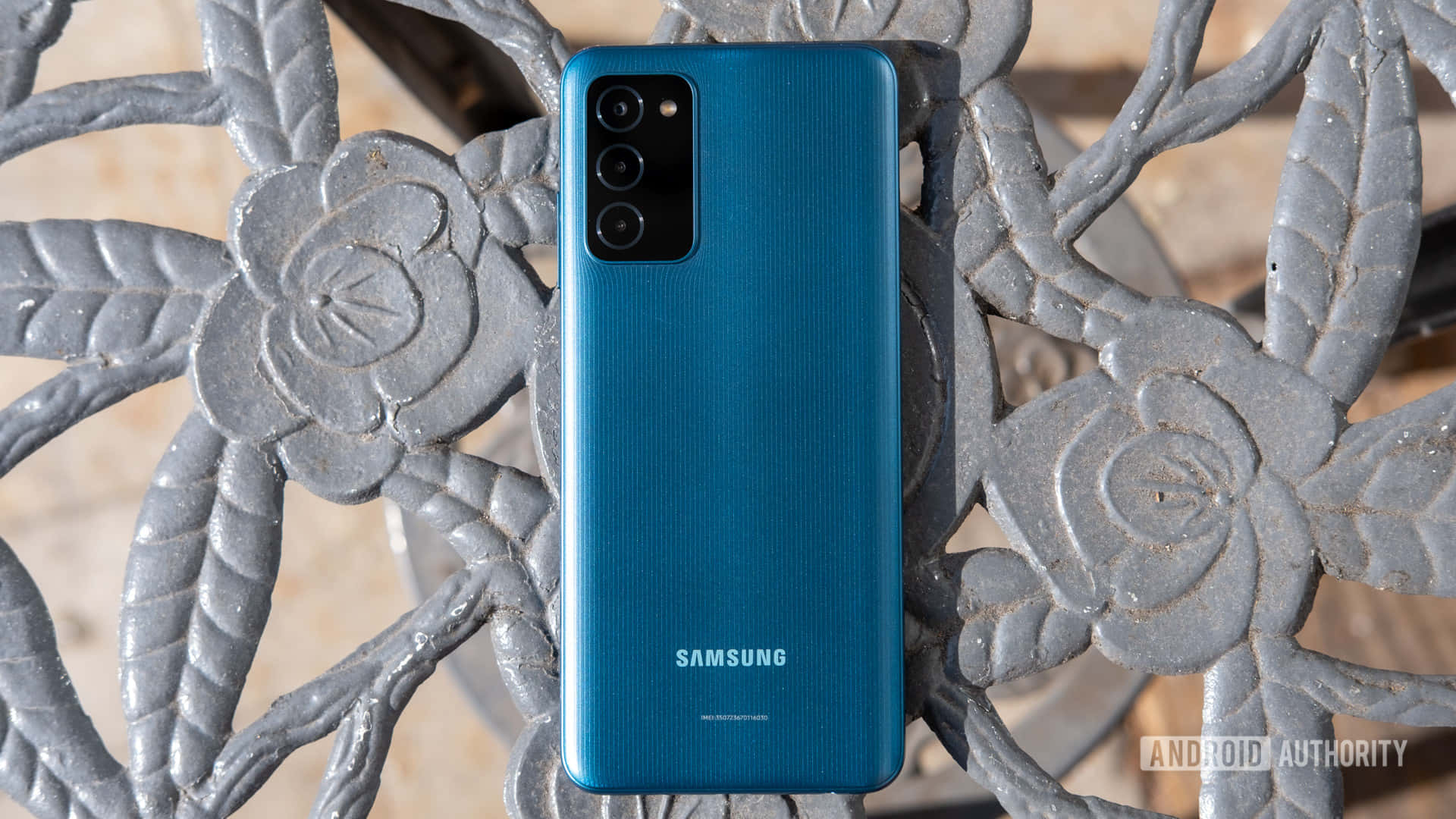 三星 桌布 壁紙 — 一部充滿活力的藍色Samsung智慧型手機直立放在一張帶有花卉圖案的華麗灰色金屬長椅上。