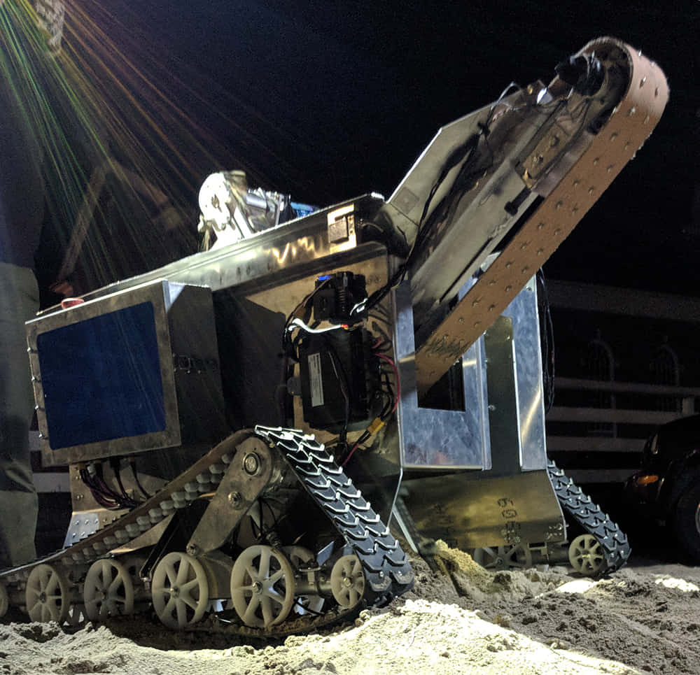 Space Mining 桌布 壁紙 — 一部堅固的履帶機器人，帶有裸露的電線和舉起的手臂，在戲劇性的夜間燈光下站在沙地上。