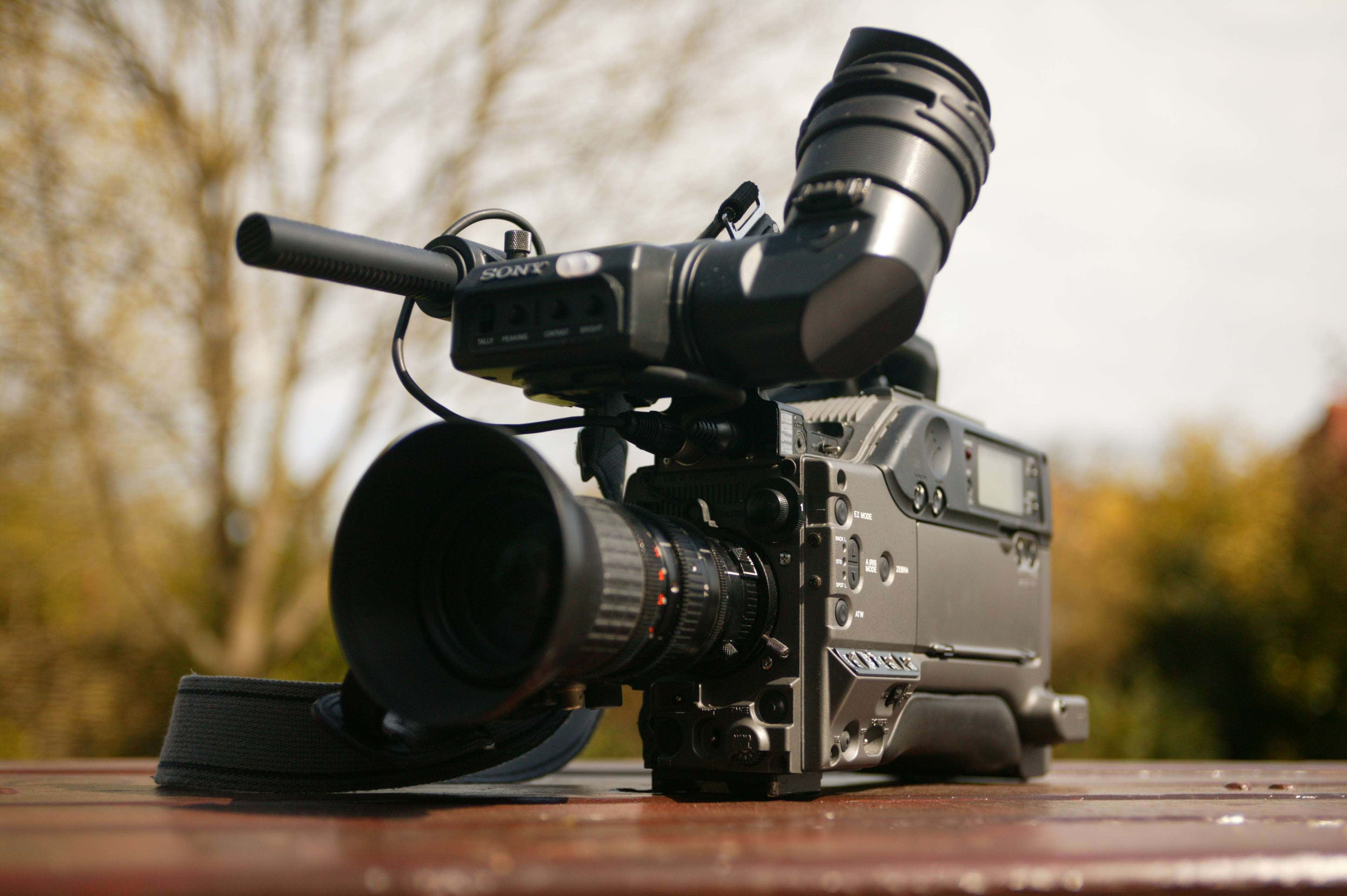 Broadcast 桌布 壁紙 — 一部專業Sony攝影機，配備大型鏡頭和麥克風，放置在戶外木質表面上，背景是模糊的秋樹。