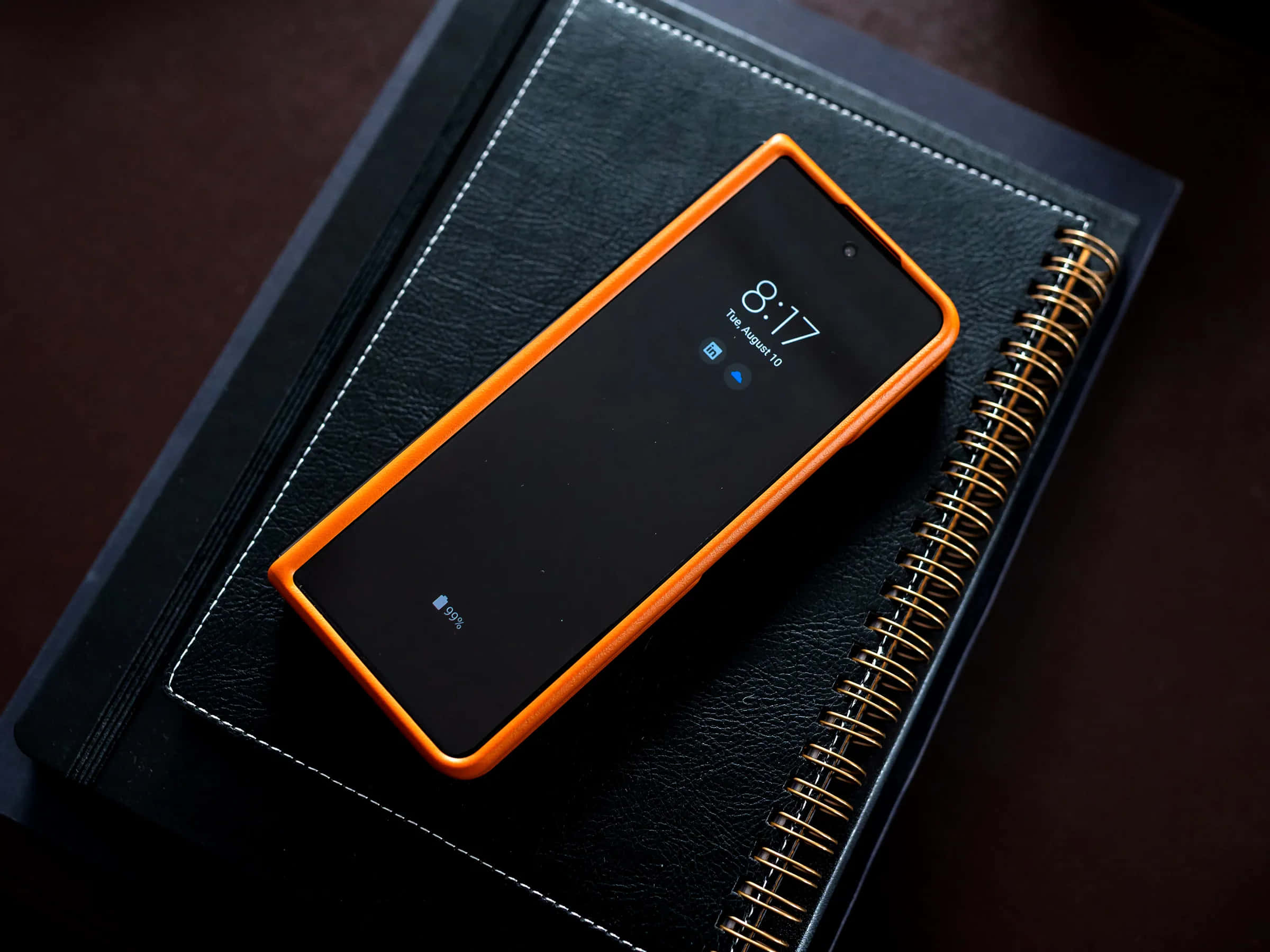 Device 桌布 壁紙 — 一部帶有亮橙色保護殼的智慧型手機放在一本黑色皮面金色螺旋裝訂的筆記本上。