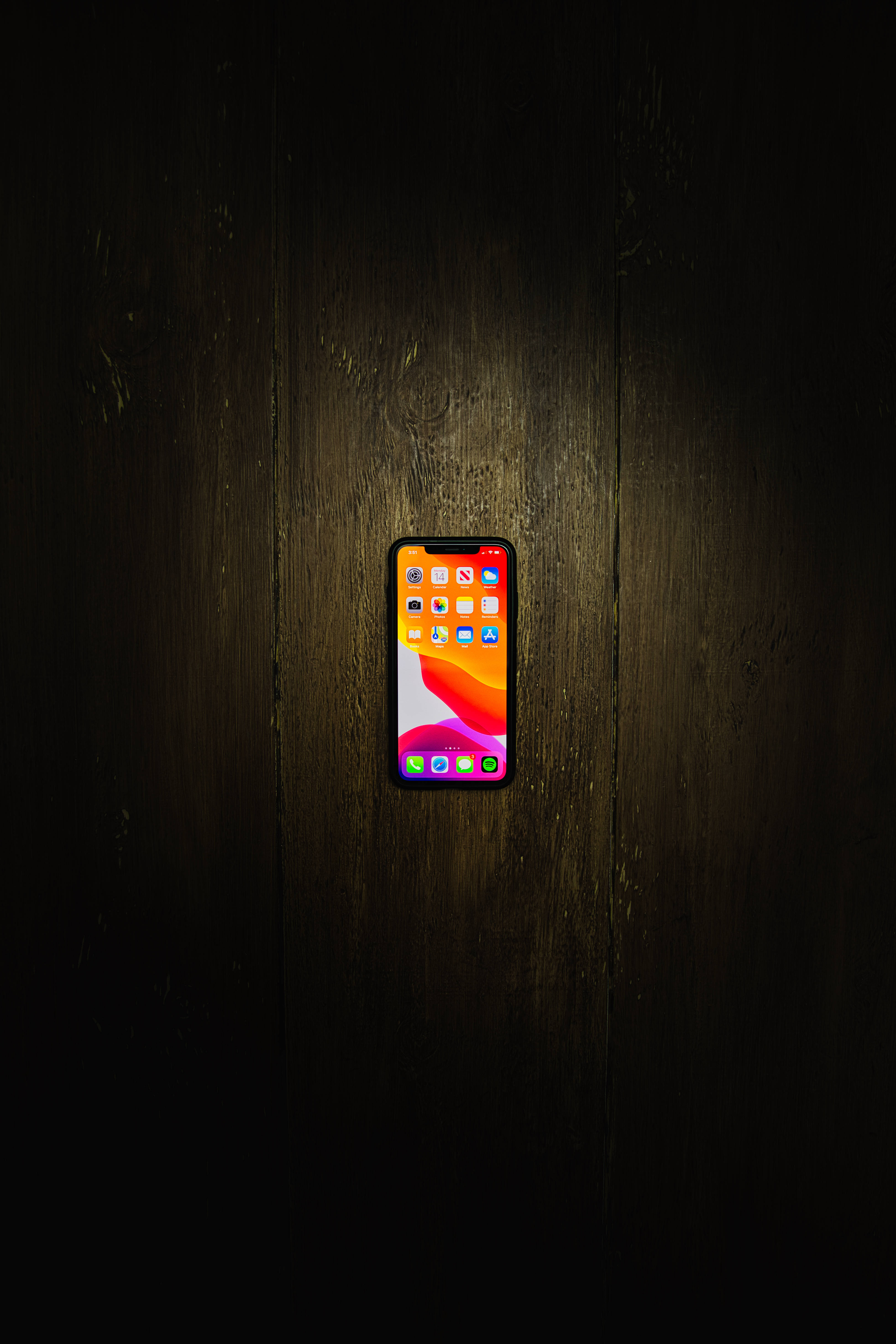 iPhone Xs Max 桌布 壁紙 — 一部帶有彩色應用程式網格的智慧型手機在聚焦照明下，在深色木質表面上柔和發光。