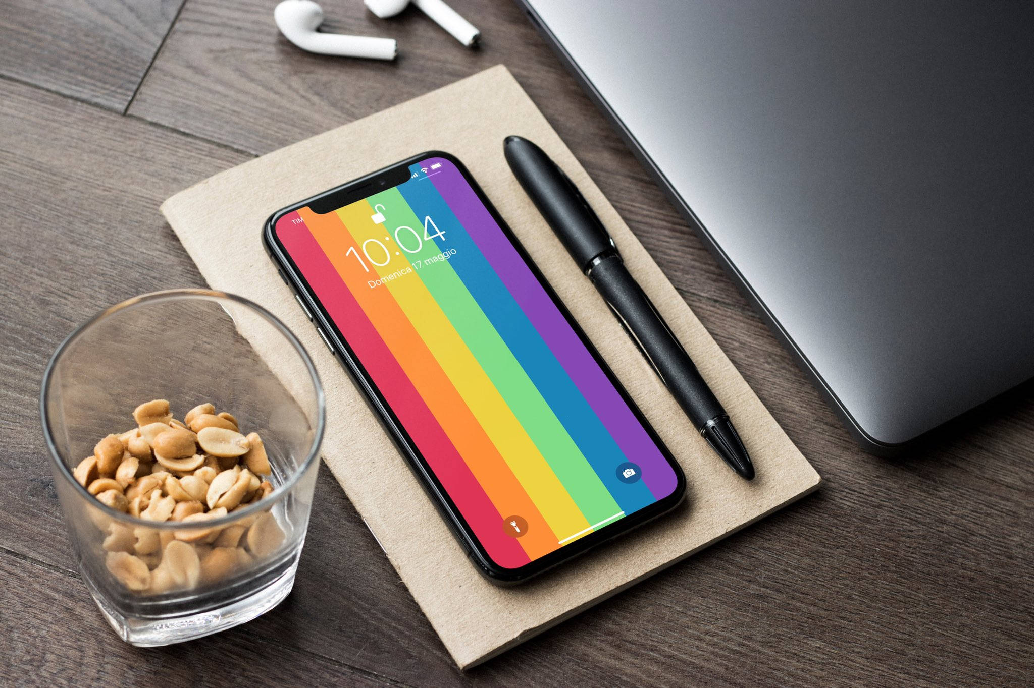 Colorful Aesthetic 桌布 壁紙 — 一部帶有彩虹桌布的智慧型手機放在木桌上，旁邊有一杯花生、一支筆和一台筆記型電腦。