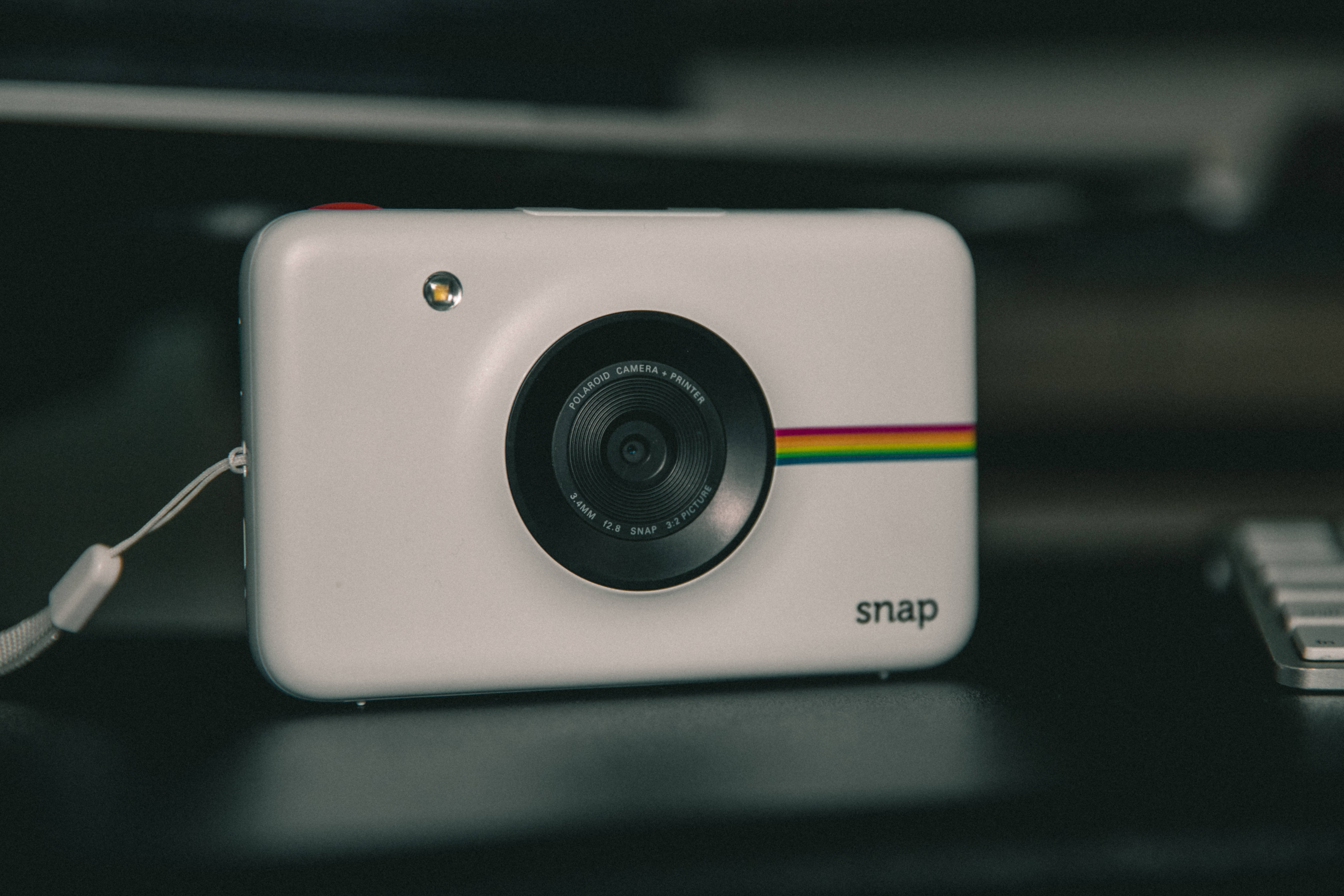 Webcam 桌布 壁紙 — 一部帶有彩虹條紋的白色Polaroid Snap相機放在深色桌子上，旁邊有一個鍵盤。