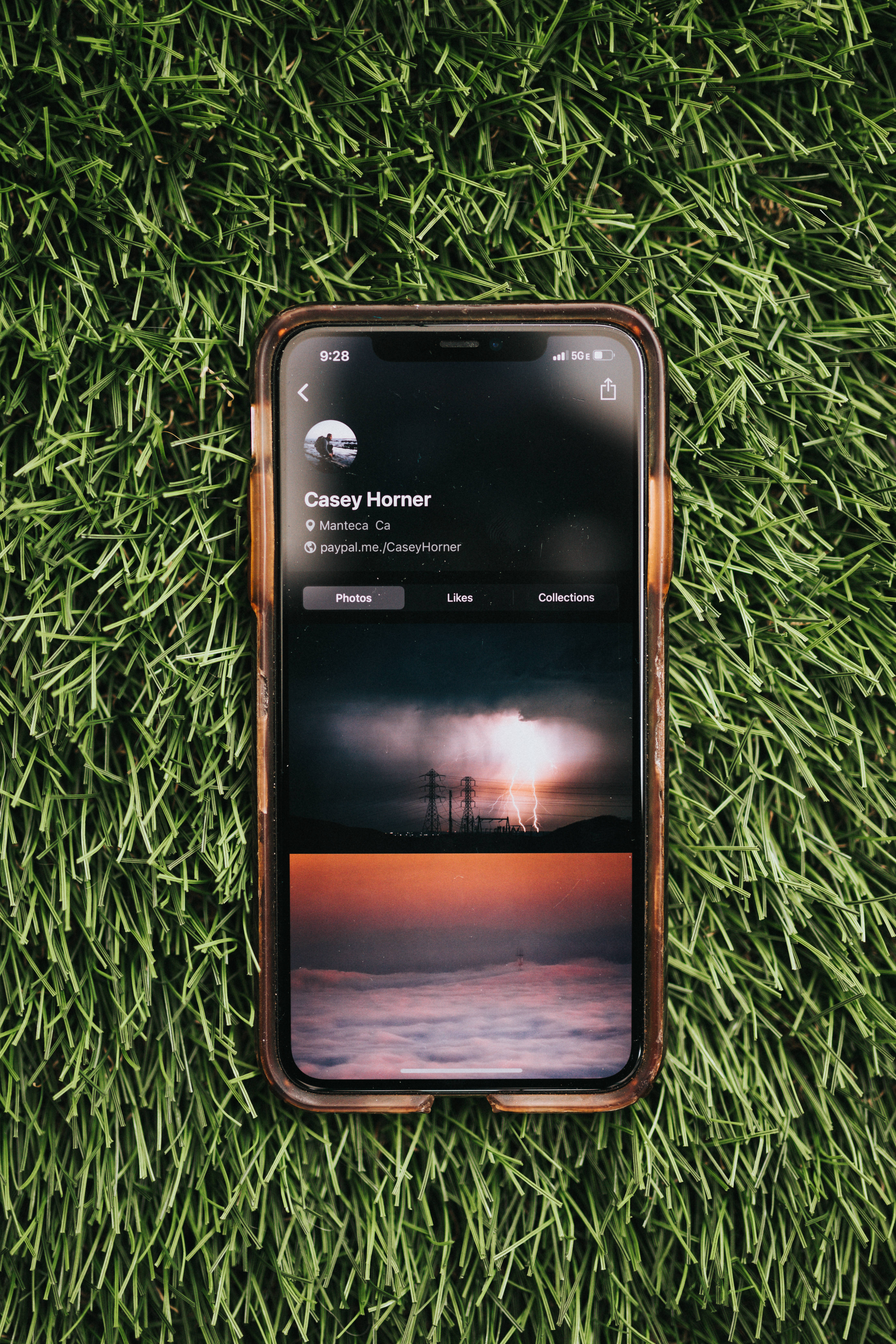 iOS 桌布 壁紙 — 一部帶有棕色保護殼的智慧型手機放在鮮綠色的草地上，顯示一個帶有暴風雨和日落照片的社交媒體個人資料。
