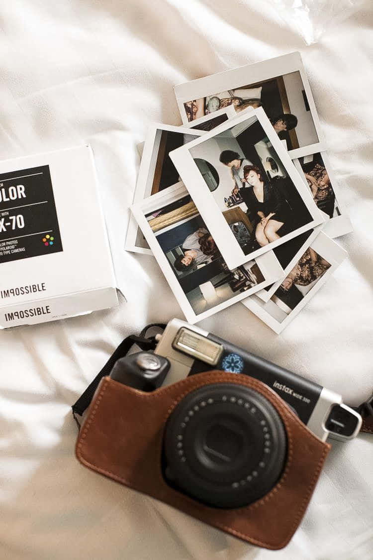 Aesthetic Polaroid 桌布 壁紙 — 一部帶有棕色皮革保護套的復古Instax相機，旁邊散落著即時照片和底片盒，放在柔軟的白色布料上。