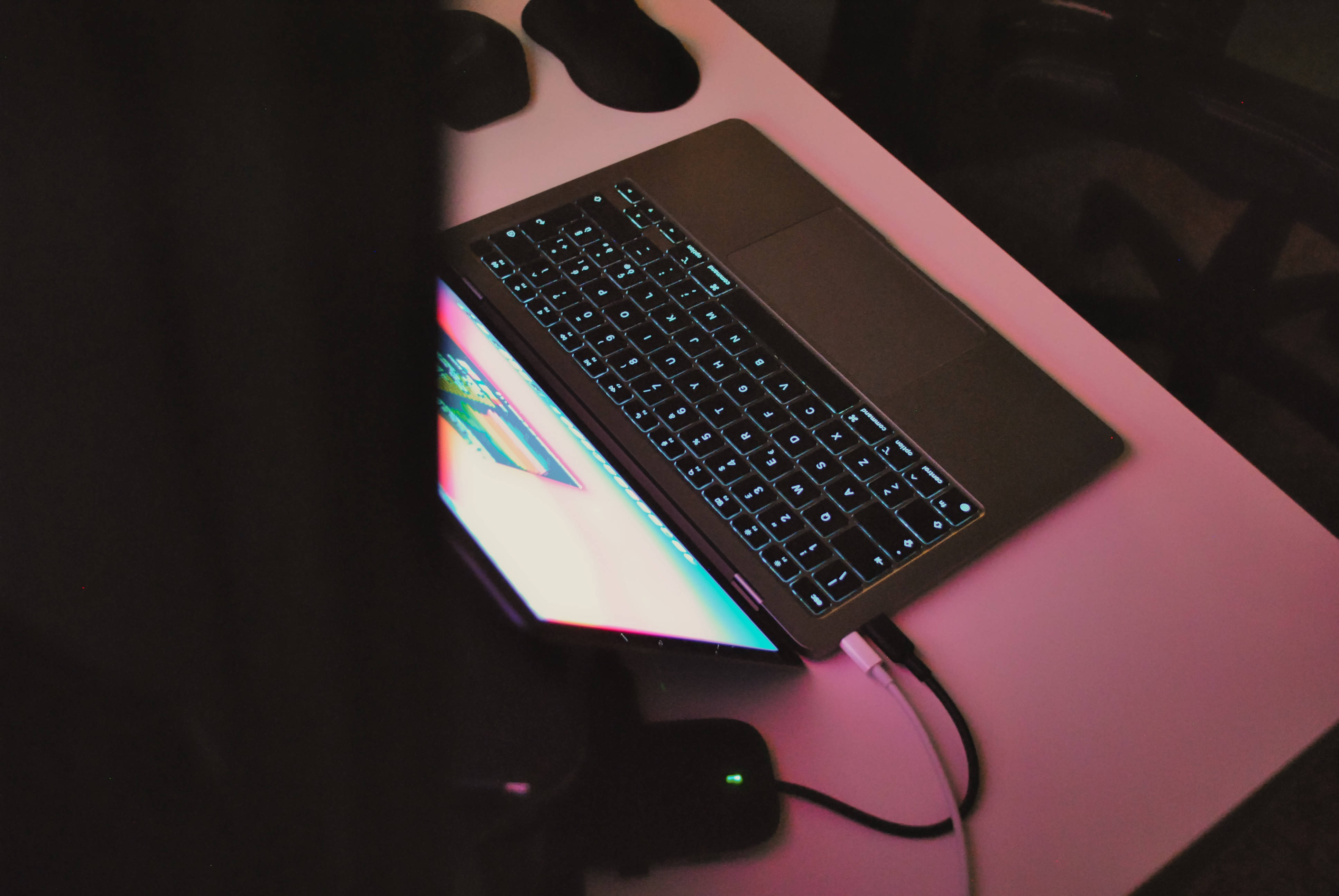Macbook Air 2020 桌布 壁紙 — 一部帶有發光鍵盤的筆記型電腦放在一張被粉紅色燈光籠罩的桌子上，附近有色彩繽紛的螢幕和週邊設備。