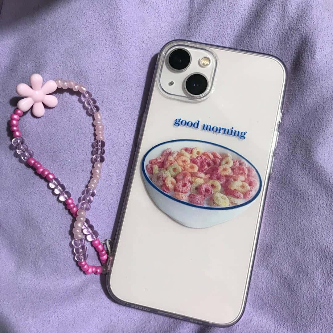 可愛 iPhone 桌布 桌布 壁紙 — 一部帶有穀物碗設計和「good morning」文字的手機，放在柔軟的紫色布料上，旁邊有一條粉紅色珠子腕帶。