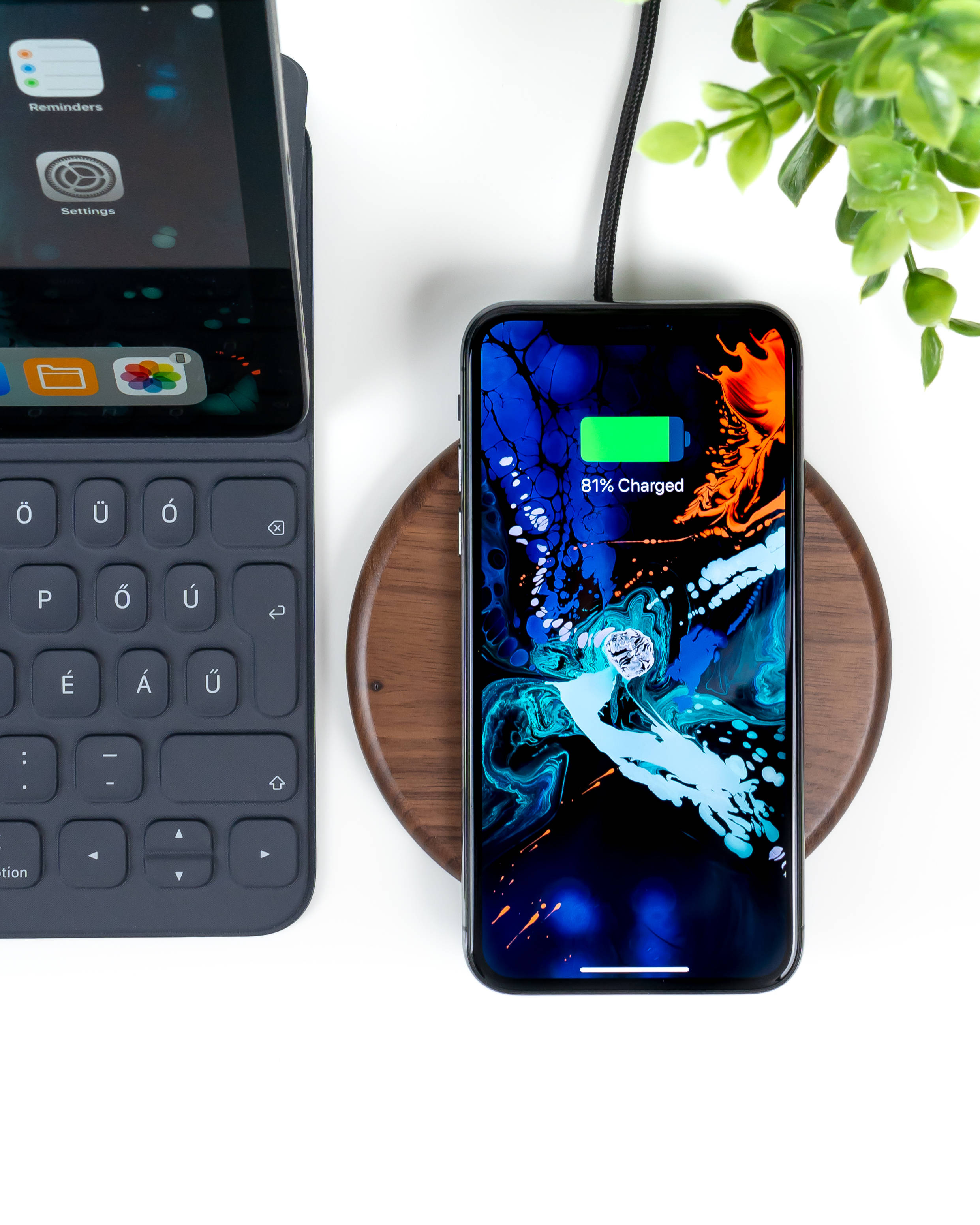 iPhone XS 桌布 壁紙 — 一部帶有鮮豔抽象桌布的智慧型手機正在木製充電板上無線充電，旁邊有鍵盤和綠色植物。