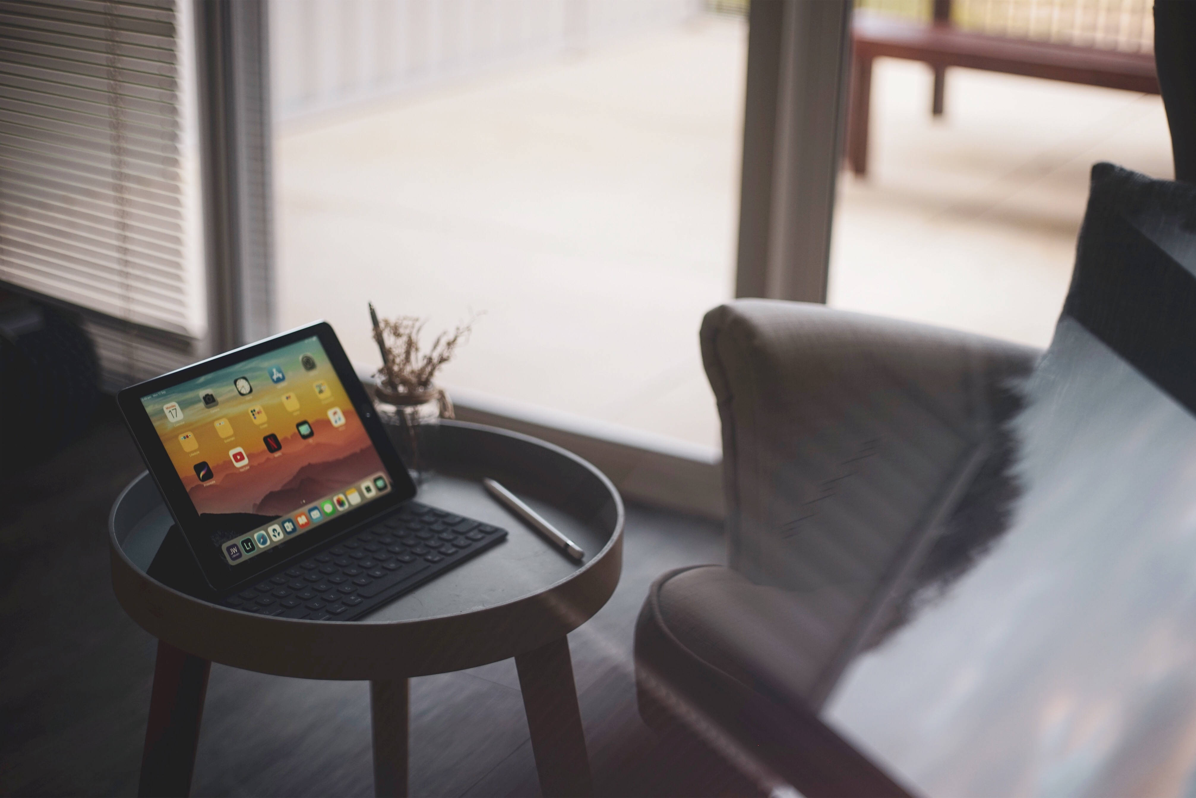 Apple iPad Pro 桌布 壁紙 — 一部帶鍵盤的平板電腦放在木製邊桌上，旁邊是一張舒適的扶手椅，靠近帶百葉窗的玻璃門。