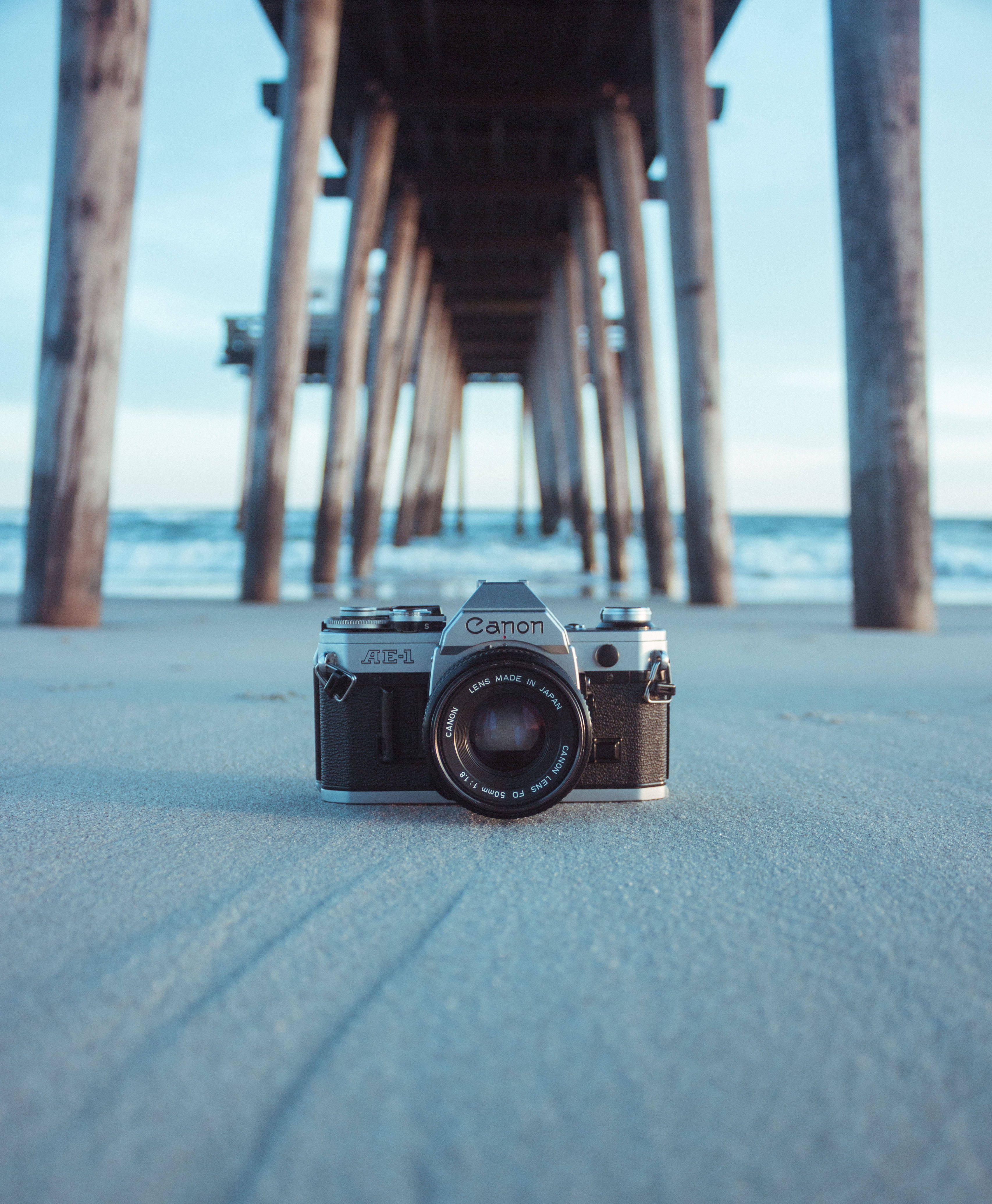 相機 桌布 壁紙 — 一部復古 Canon AE-1 底片相機放置在沙灘上，位於木製碼頭樁柱下方，背景有海浪。