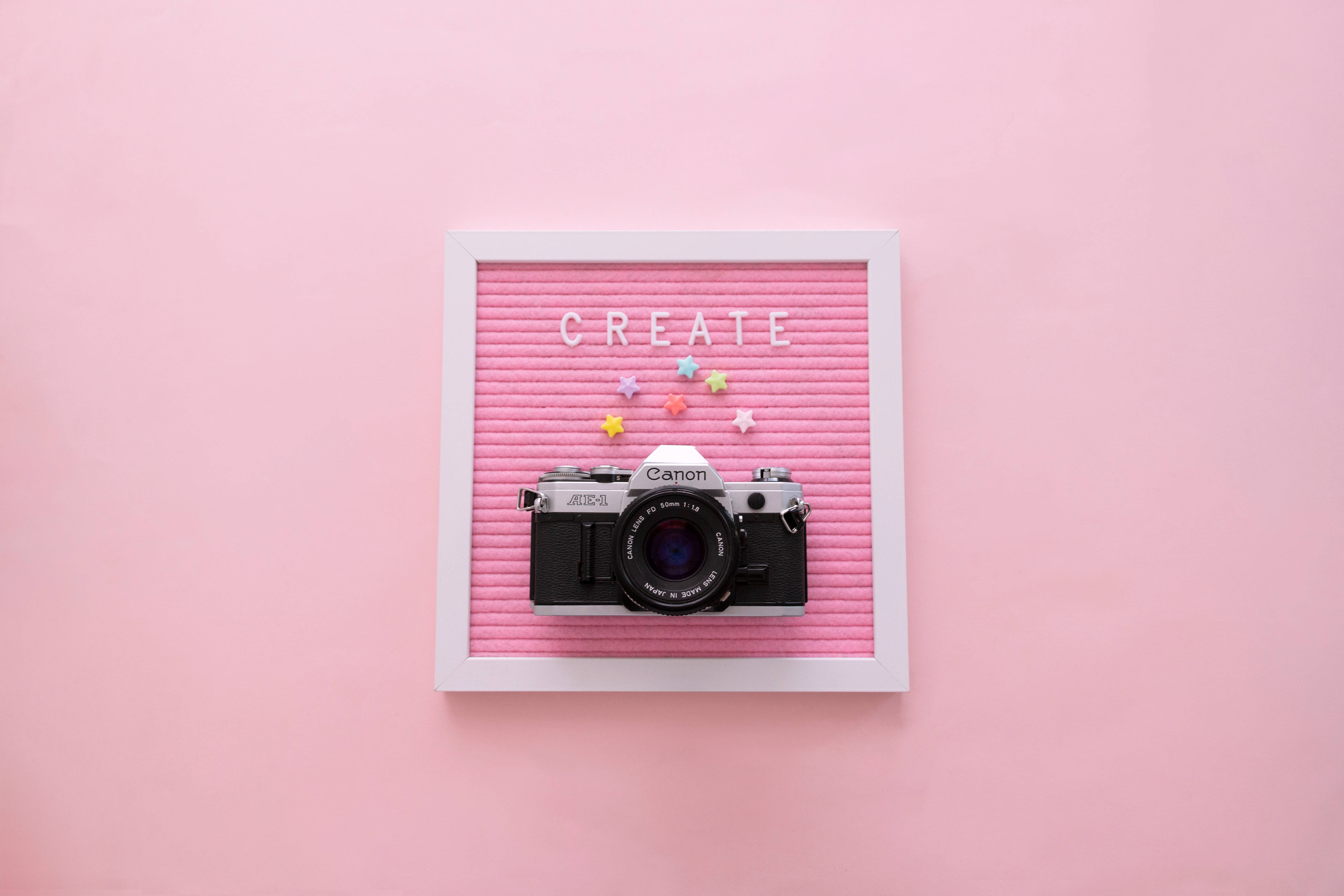 girly pink aesthetic 桌布 壁紙 — 一部復古 Canon 相機放在白色框邊的粉紅色氈板上，上面用字母和彩色星星拼出 CREATE 這個詞。
