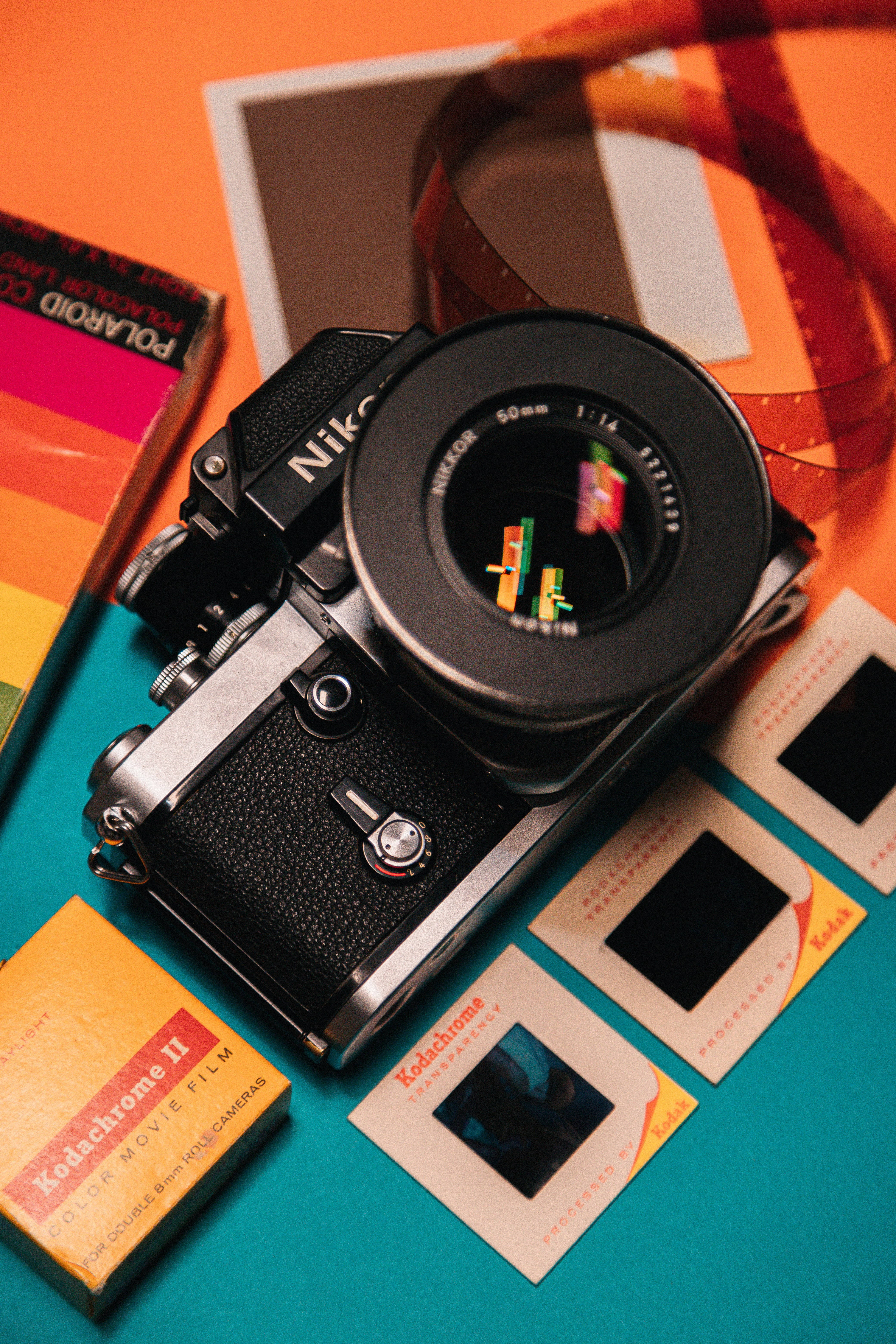 橘色 桌布 壁紙 — 一部復古 Nikon 底片相機放置在充滿活力的青綠色和橙色表面上，周圍環繞著 Kodachrome 底片盒和幻燈片。