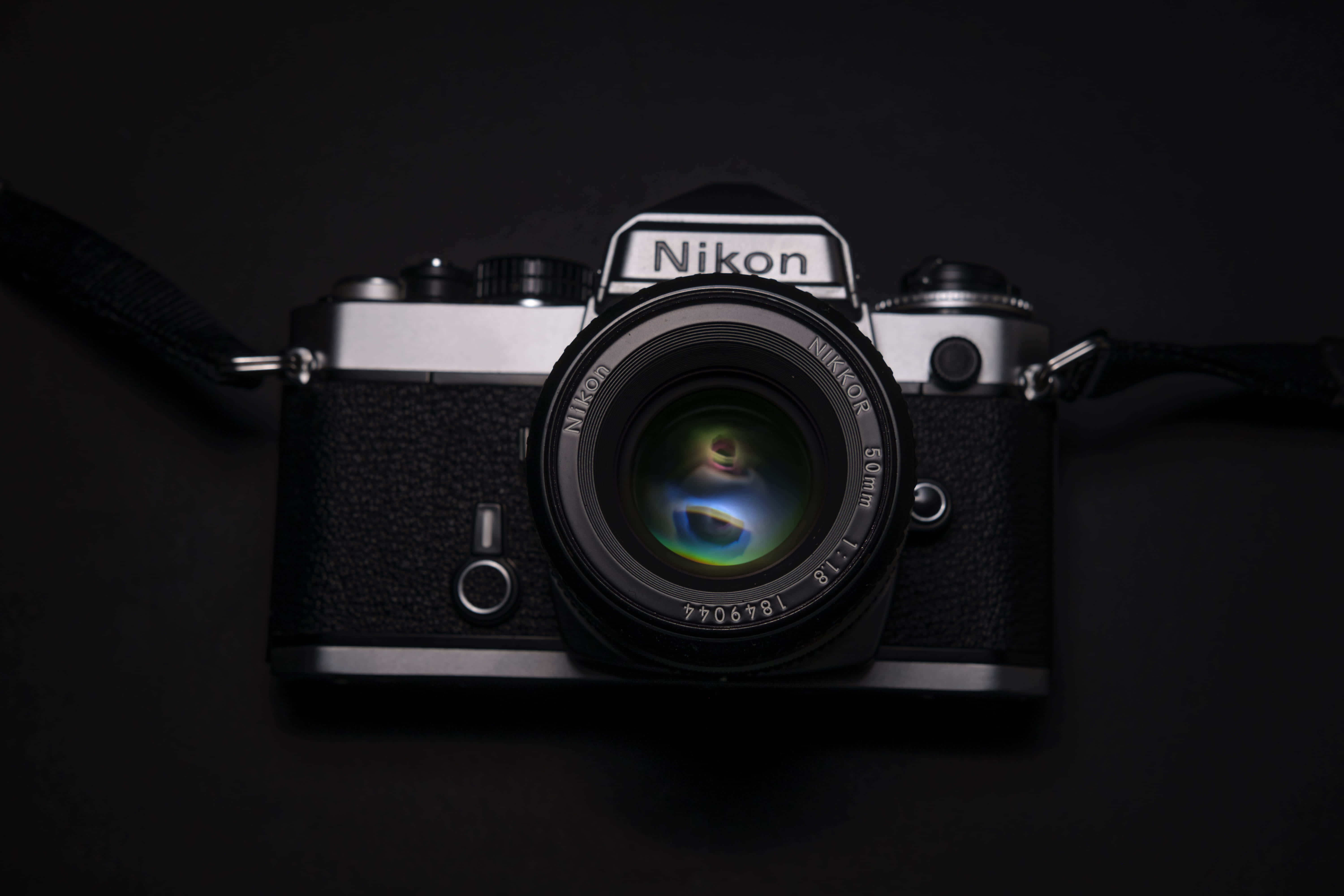 Dslr 桌布 壁紙 — 一部復古 Nikon 底片相機配備 50mm 鏡頭，放置在黑色表面上，鏡頭在昏暗的攝影棚中反射出鮮豔色彩。