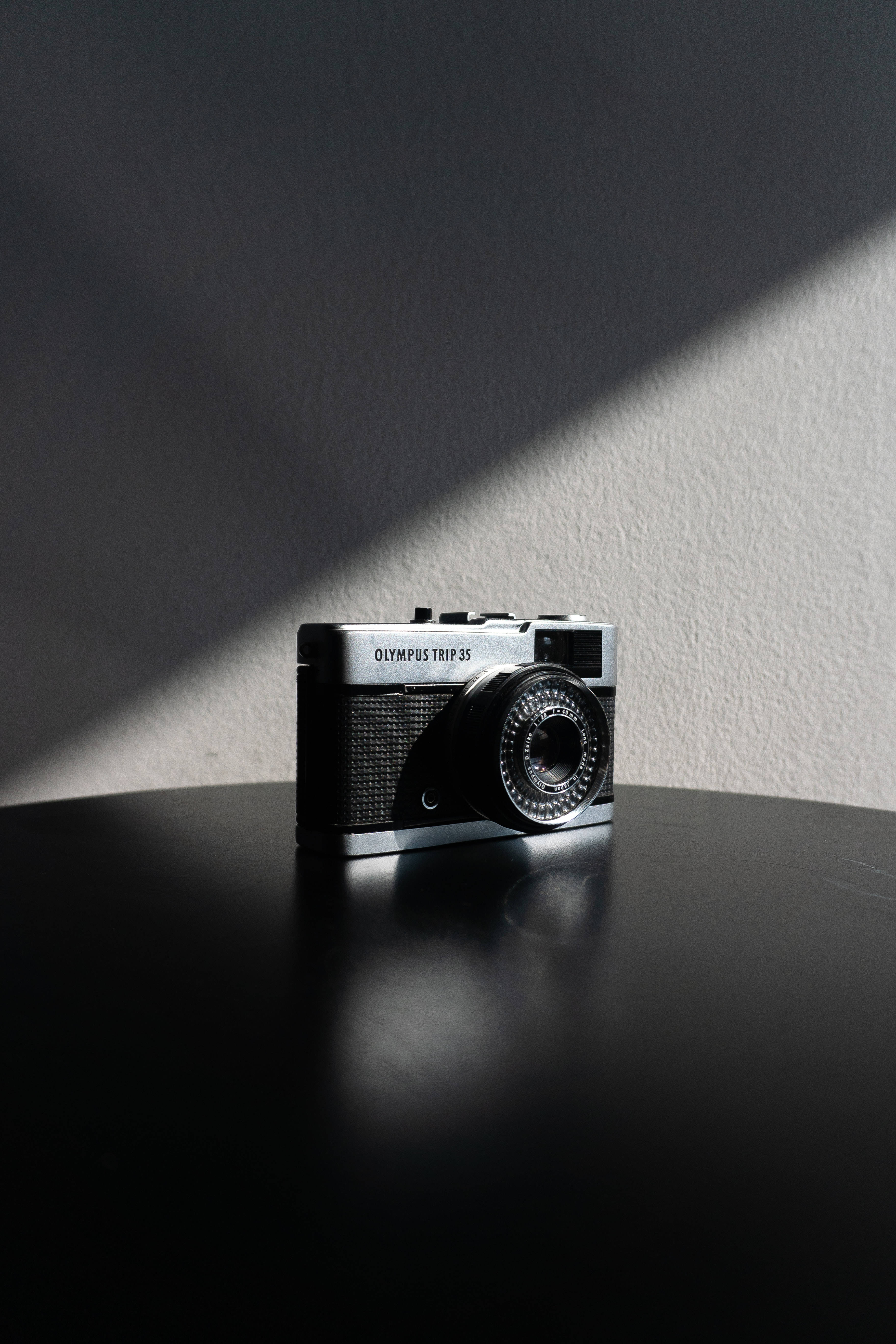 預科生美學 桌布 壁紙 — 一部復古Olympus Trip 35相機放置在黑色表面上，沐浴在戲劇性的對角陽光中，背景是紋理牆壁。
