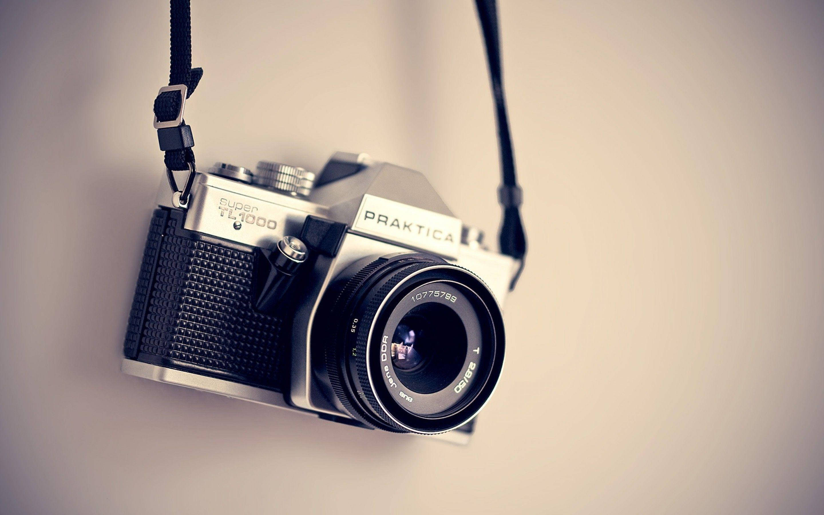 相機 桌布 壁紙 — 一部復古Praktica TL1000底片相機用掛帶掛在柔軟的米色牆上，帶有溫暖的燈光。
