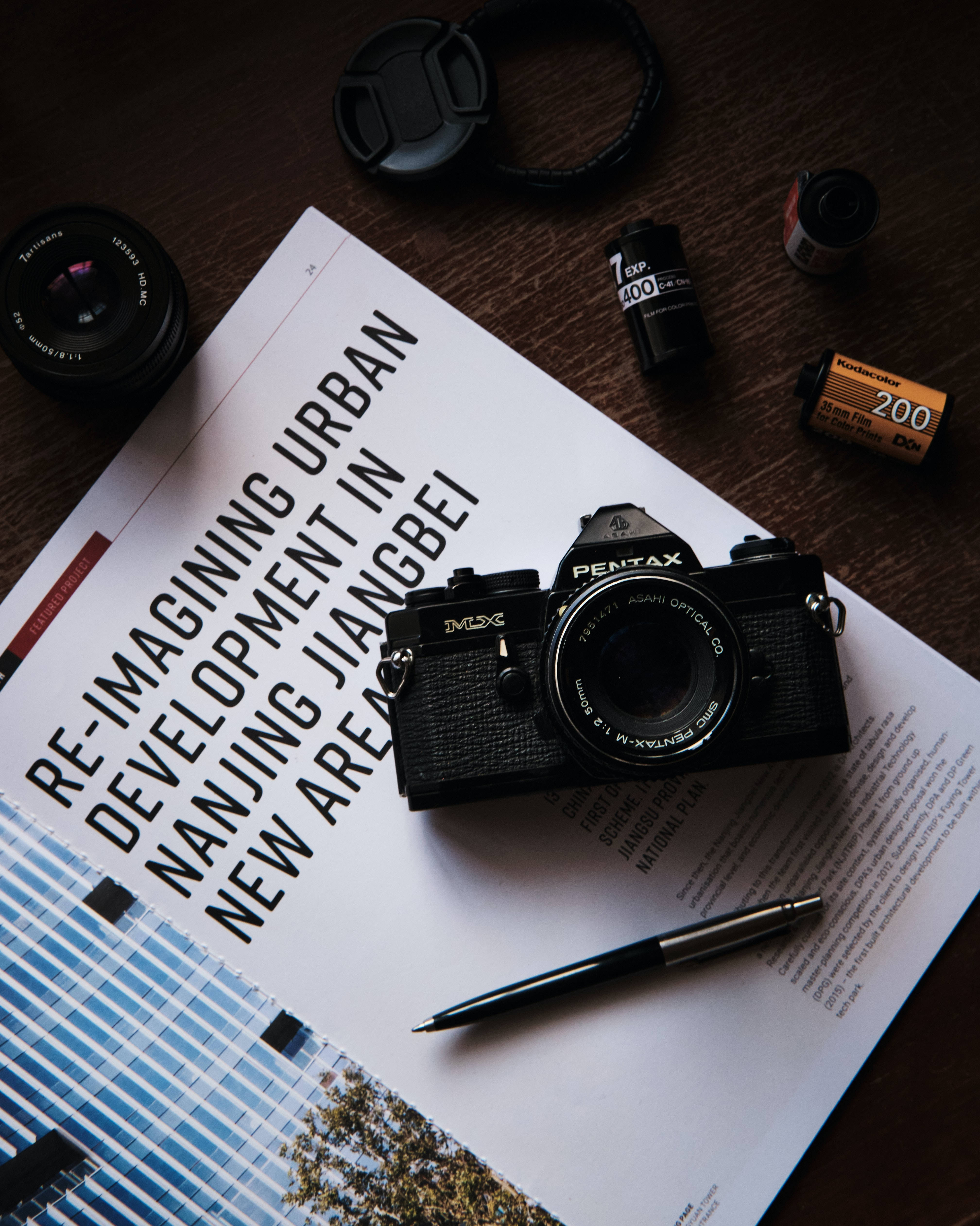 Magazine 桌布 壁紙 — 一部復古黑色Pentax底片相機放在一本關於城市發展的雜誌上，周圍有鏡頭和底片卷在木質表面上。