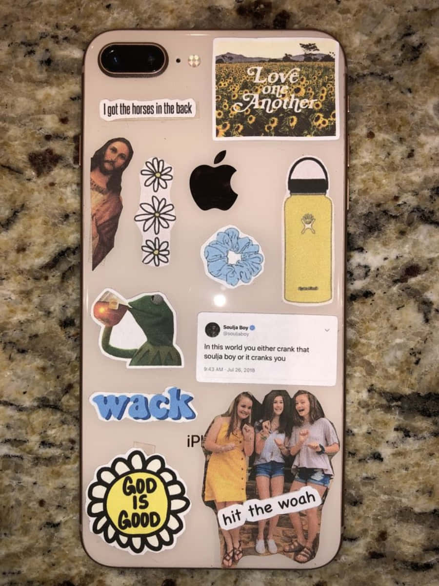 美學 iPhone 壁紙 桌布 壁紙 — 一部手機上貼滿了彩色貼紙，包括耶穌、向日葵、青蛙和社交媒體帖子，放在花崗岩檯面上。