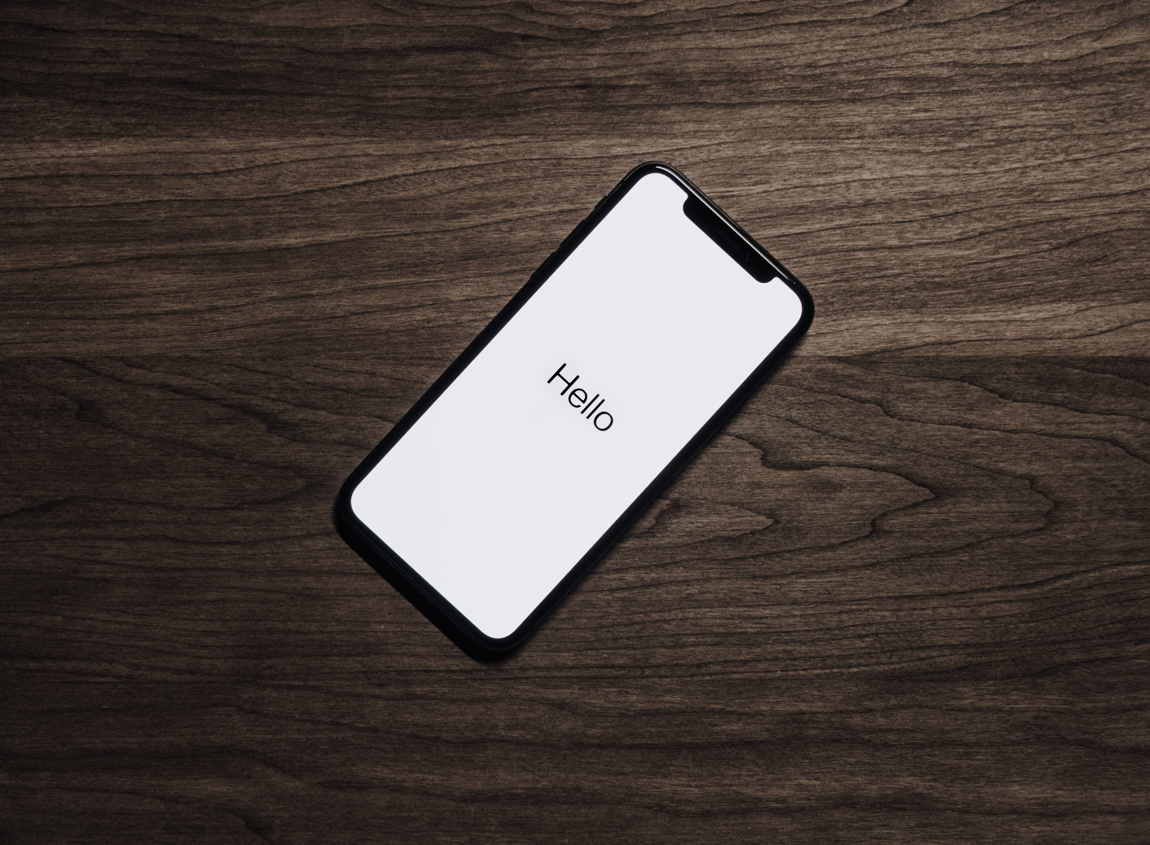 酷炫iPhone 桌布 壁紙 — 一部時尚智慧型手機，白色螢幕上顯示「hello」字樣，斜放在深色紋理木質表面上。