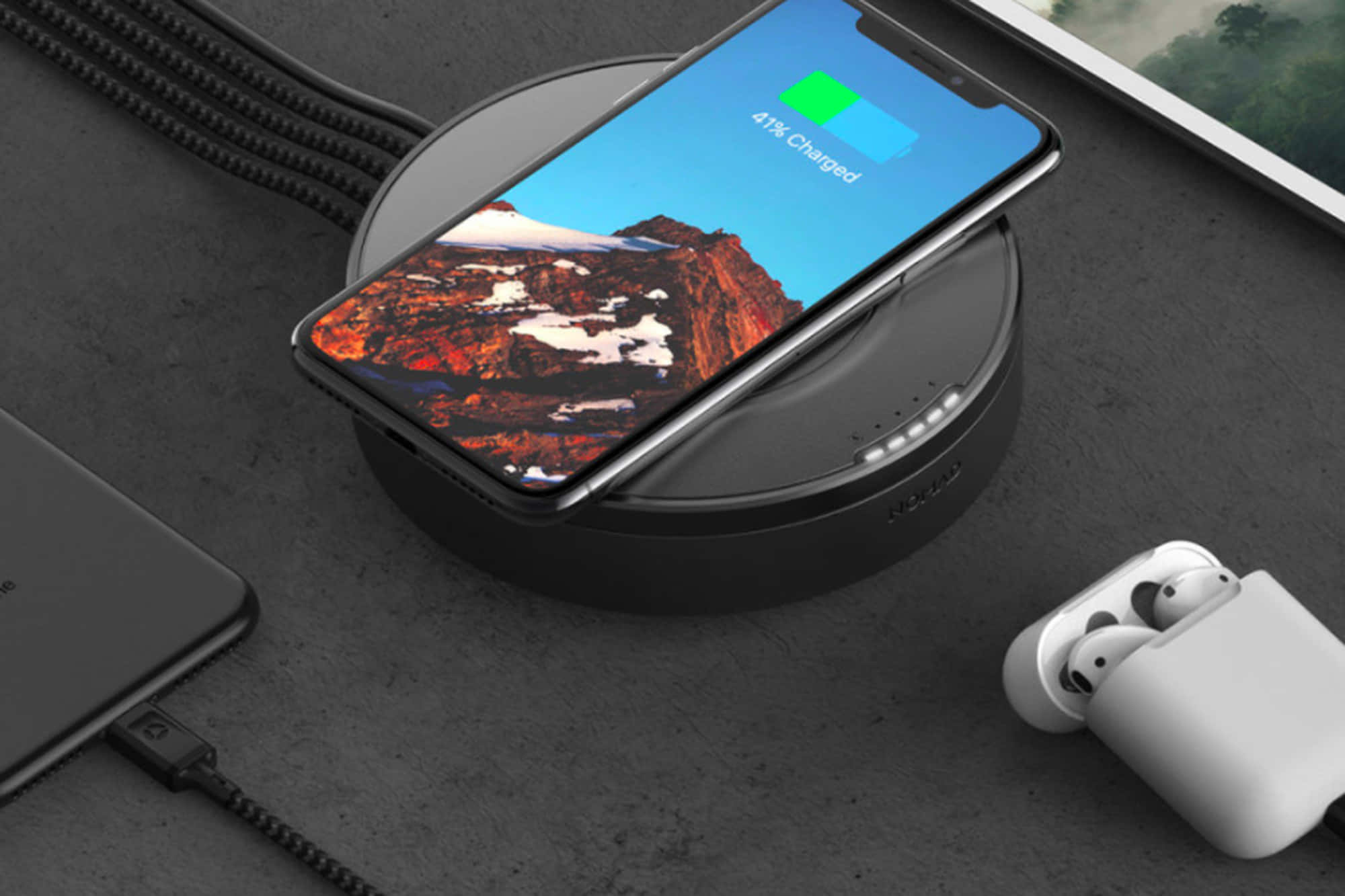 Wireless Charging 桌布 壁紙 — 一部時尚智慧型手機在顯示山景桌面的黑色充電板上無線充電，而附近的耳機和平板電腦則放在深色紋理表面上。