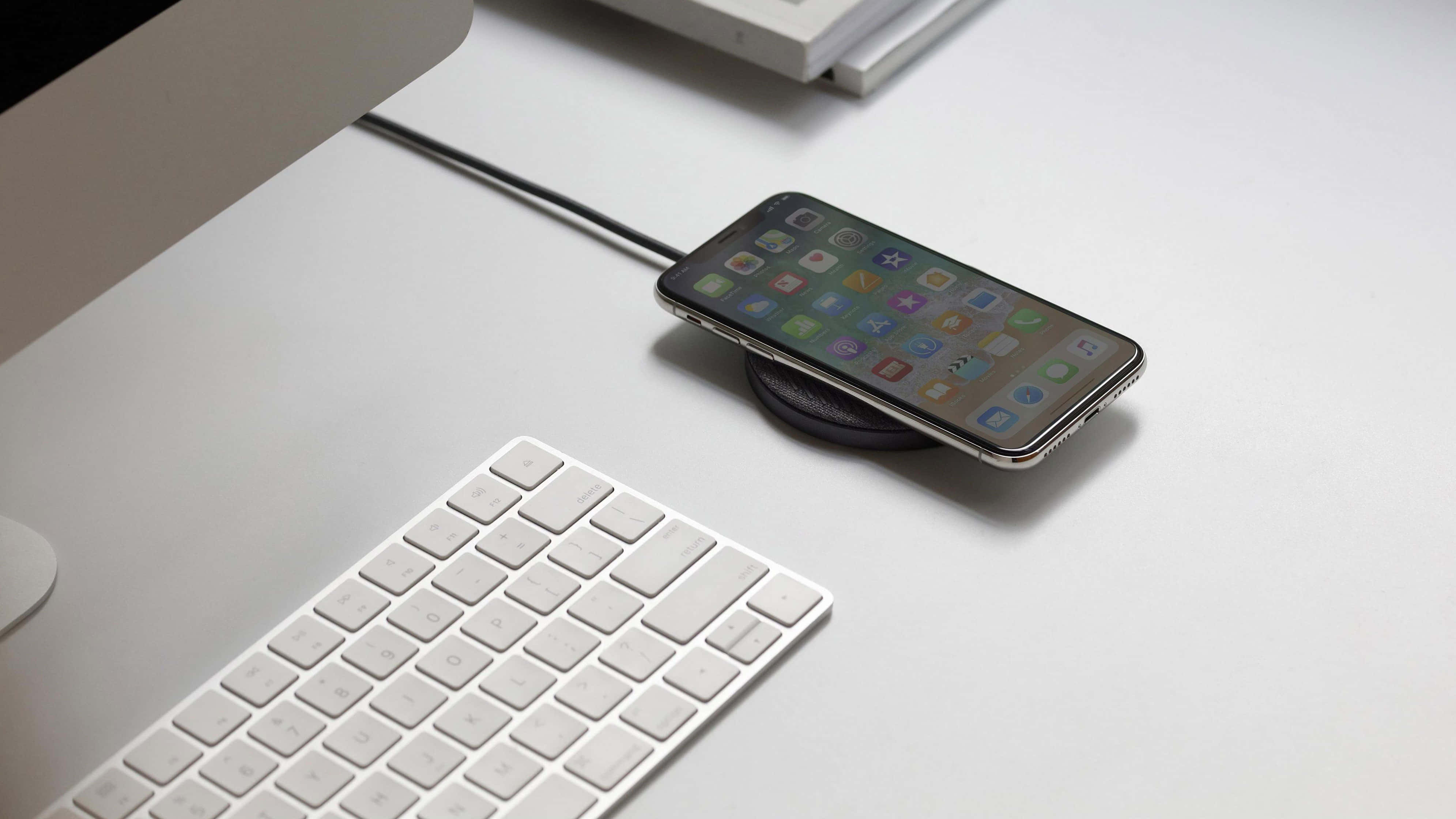 Wireless Charging 桌布 壁紙 — 一部時尚智慧型手機放在無線充電器上，旁邊是白色鍵盤和顯示器，置於簡約白色桌上。