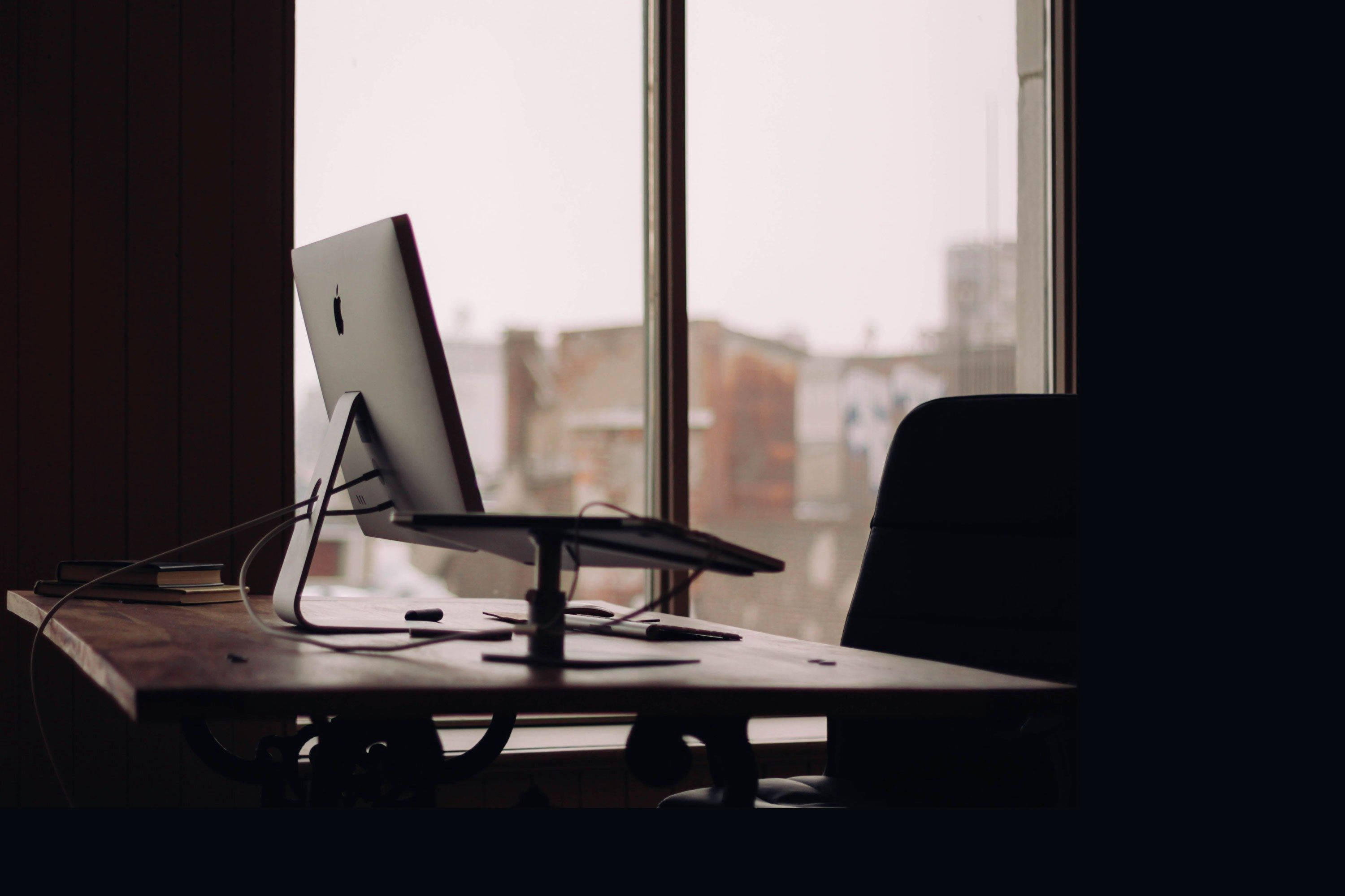 桌面 桌布 壁紙 — 一部時尚的 iMac 放在木桌上，旁邊是窗戶，窗外是柔和日光下的朦朧城市景觀。