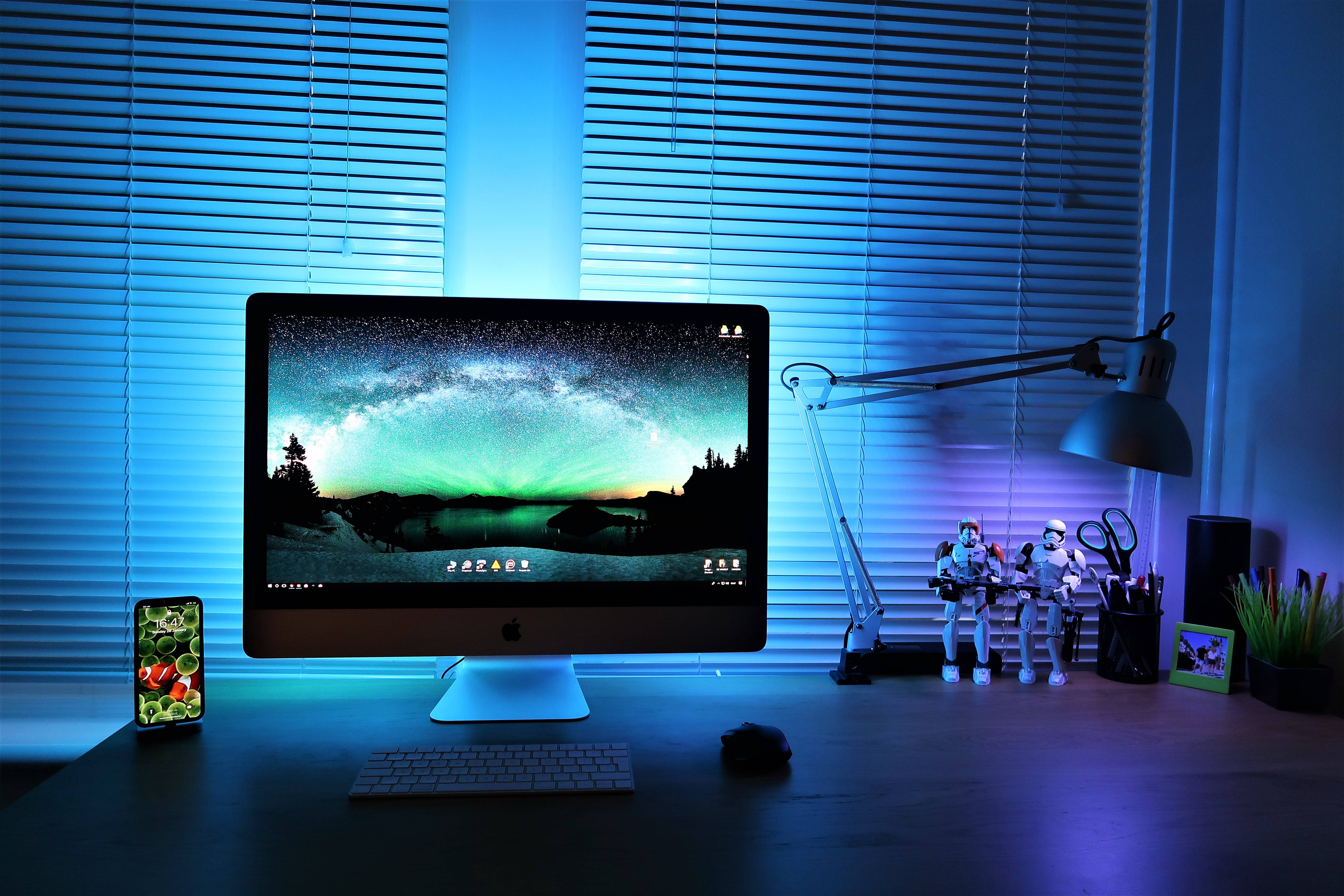 Mac 桌布 壁紙 — 一部時尚的 iMac 顯示著充滿活力的極光桌布，而發光的藍色環境光線灑滿整潔的桌面，上面有風暴兵人偶和智慧型手機。