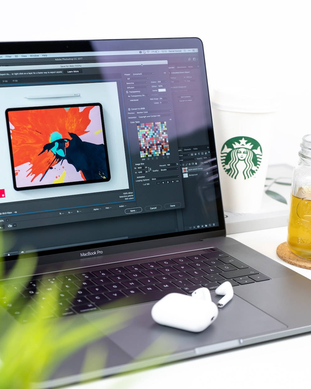 Best Laptop 桌布 壁紙 — 一部時尚的 MacBook Pro 在 Photoshop 中顯示充滿活力的數位藝術品，旁邊放著 Starbucks 杯和無線耳塞，在簡約的桌子上。