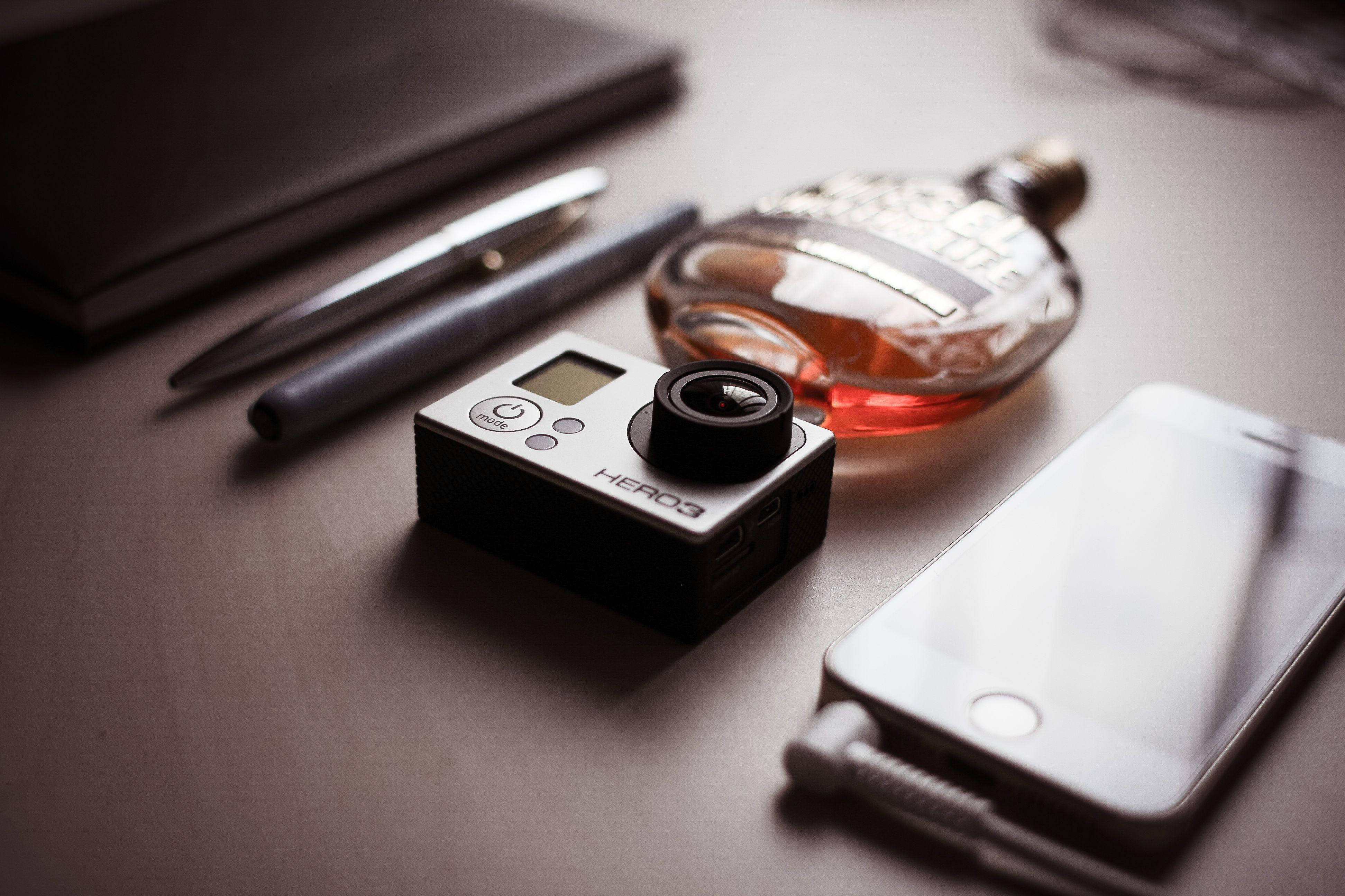 攝影 桌布 壁紙 — 一部時尚的GoPro相機放在智慧型手機威士忌酒瓶和筆旁邊，置於簡約木桌上。