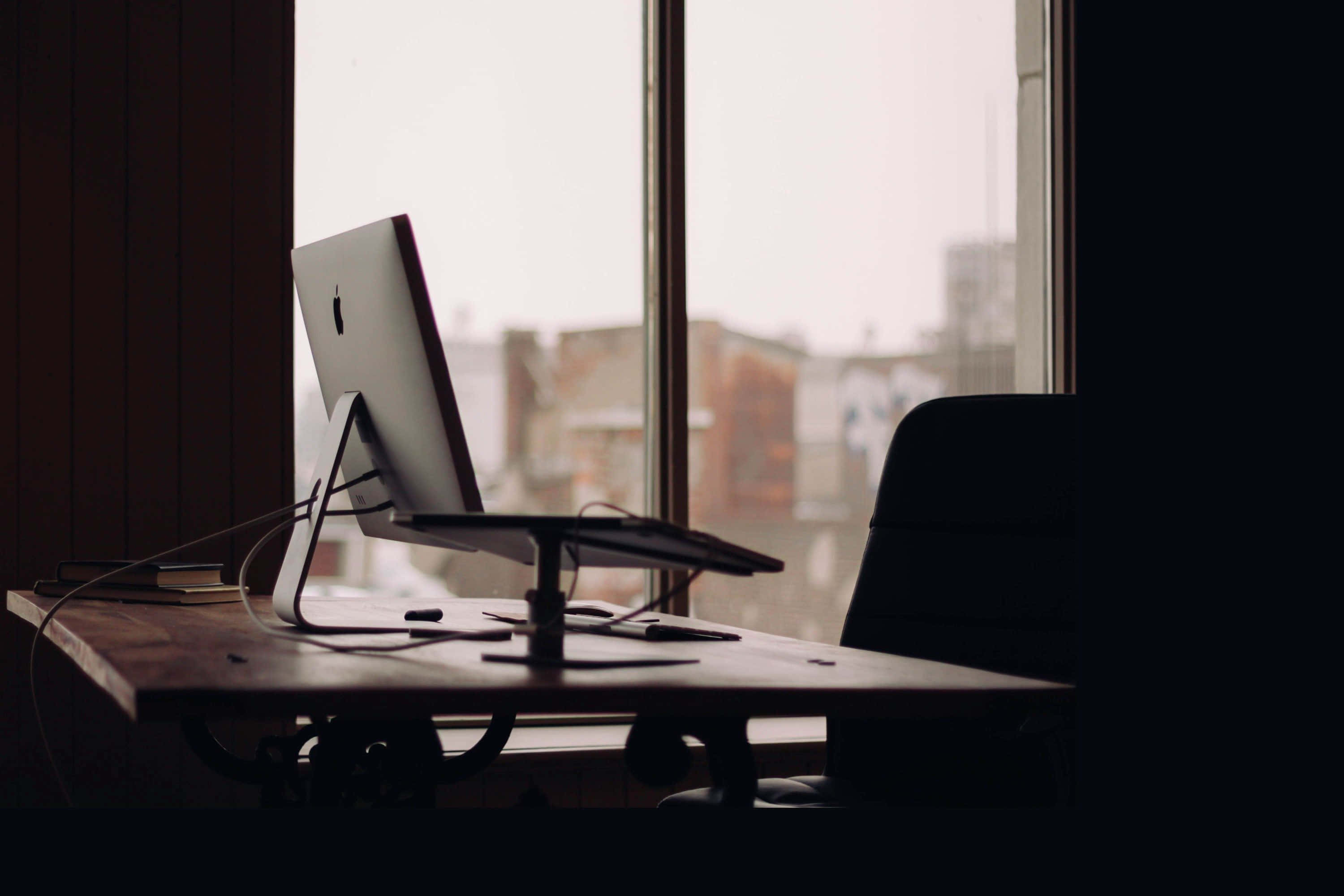 Office Desk 桌布 壁紙 — 一部時尚的iMac放在木桌上，房間燈光昏暗，大窗戶外是模糊的城市景觀。