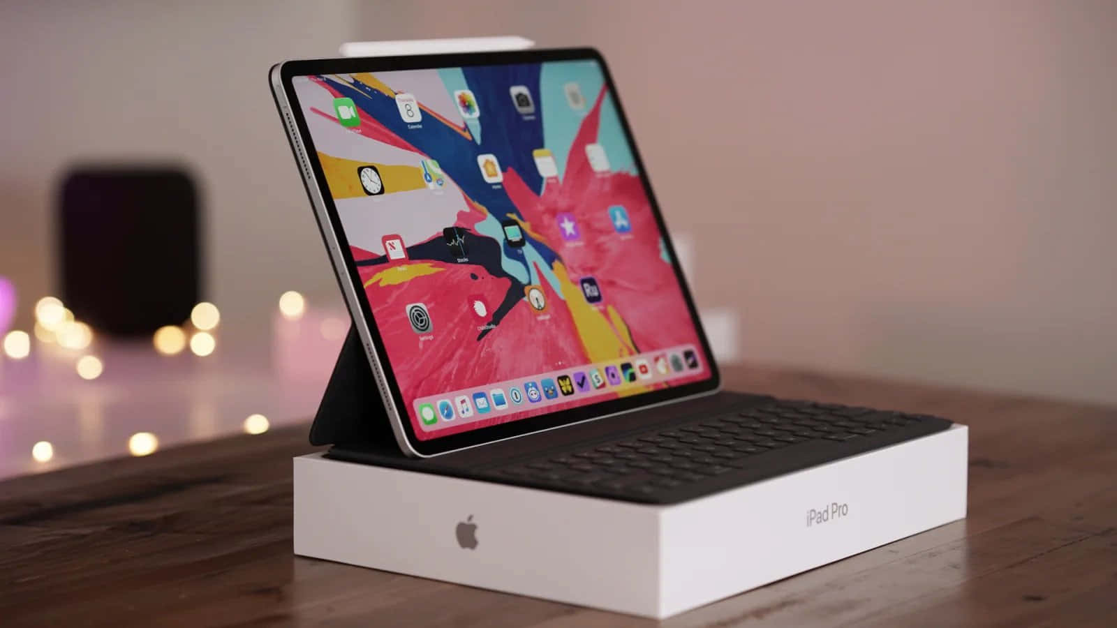 iPad 桌布 壁紙 — 一部時尚的iPad Pro搭配鍵盤保護套，放在木桌上的白色包裝盒上，發光的螢幕在溫暖的散景燈光背景下顯示色彩繽紛的應用程式。