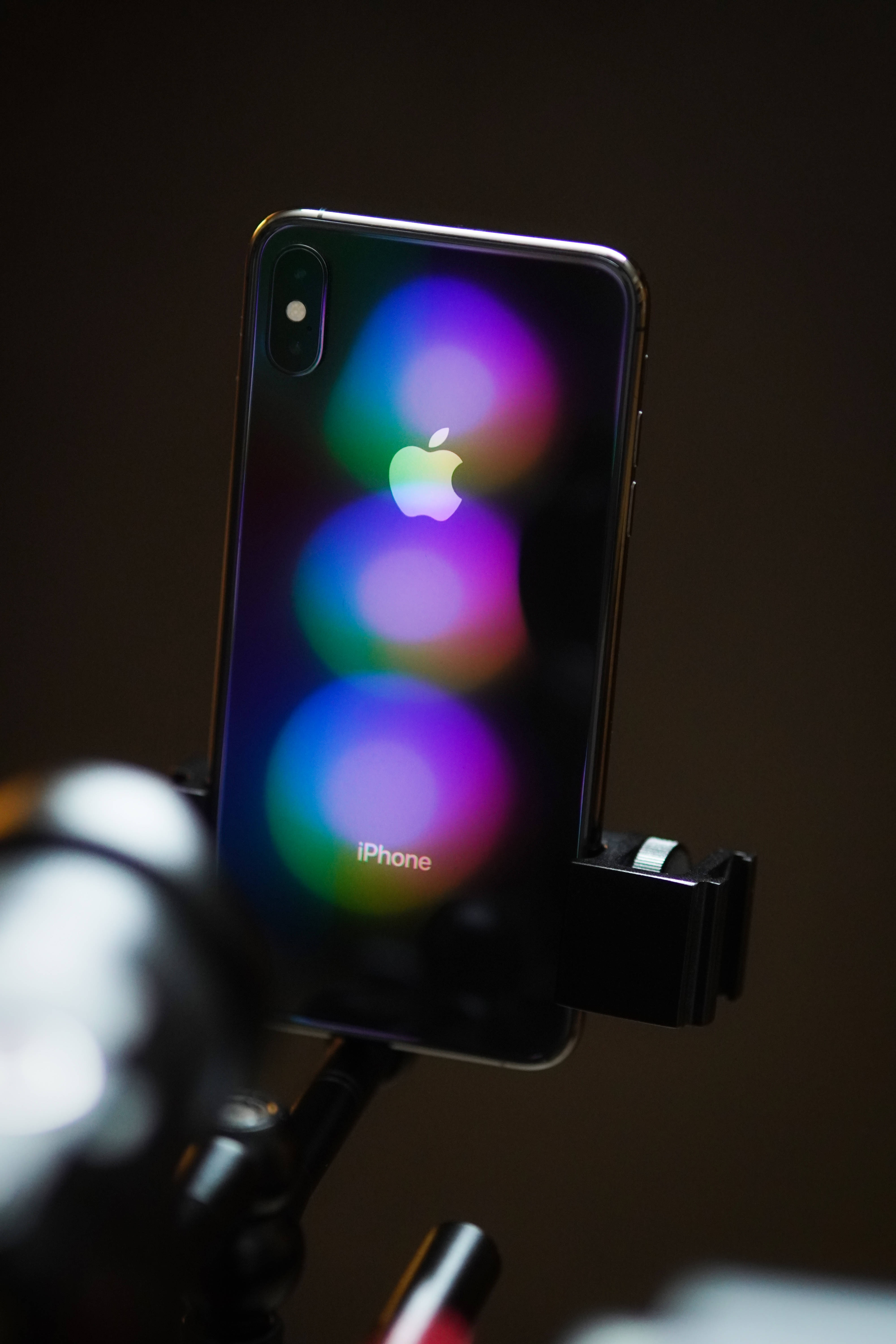 iPhone Xs Max 桌布 壁紙 — 一部時尚的iPhone X安裝在三腳架上，其反光背面後方有鮮豔的散景燈光閃爍。