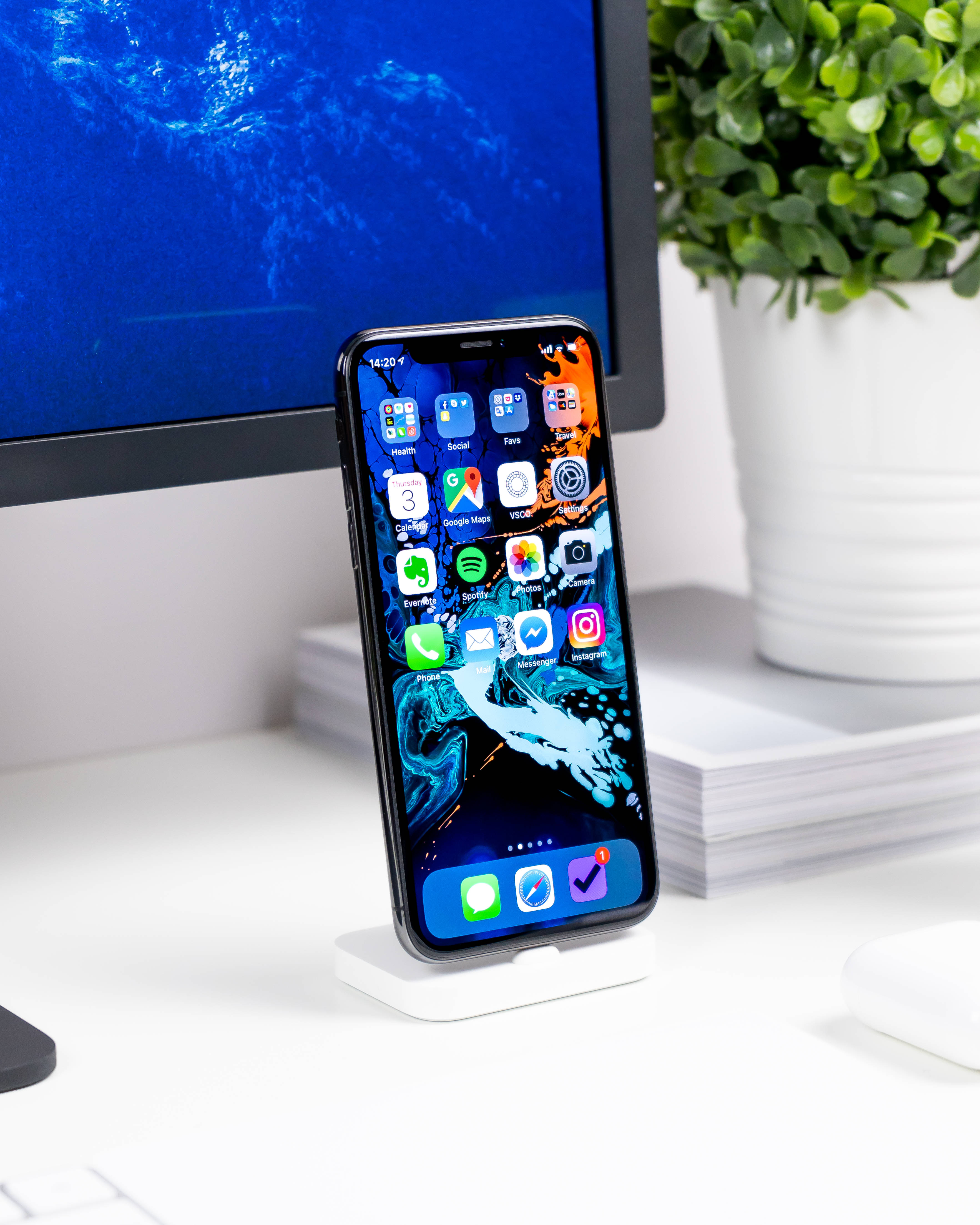 Simple iPhone 桌布 壁紙 — 一部時尚的智慧型手機帶有鮮豔的抽象桌布，直立放在白色桌子上，旁邊是顯示器和盆栽植物。