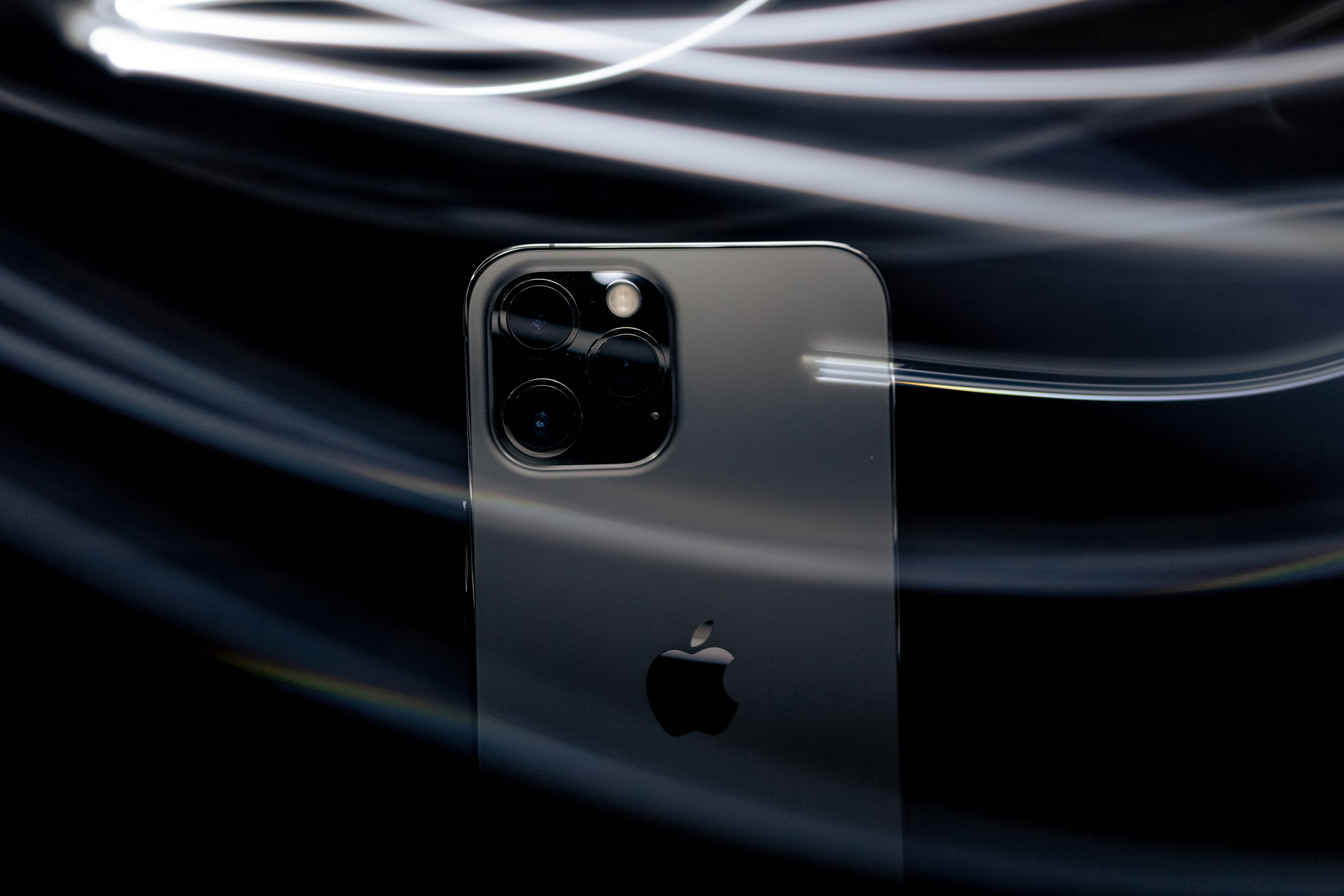 dope iPhone 桌布 壁紙 — 一部時尚的深灰色 iPhone 配備三鏡頭陣列，在黑色背景上與明亮的白色光軌動態漩渦形成對比。