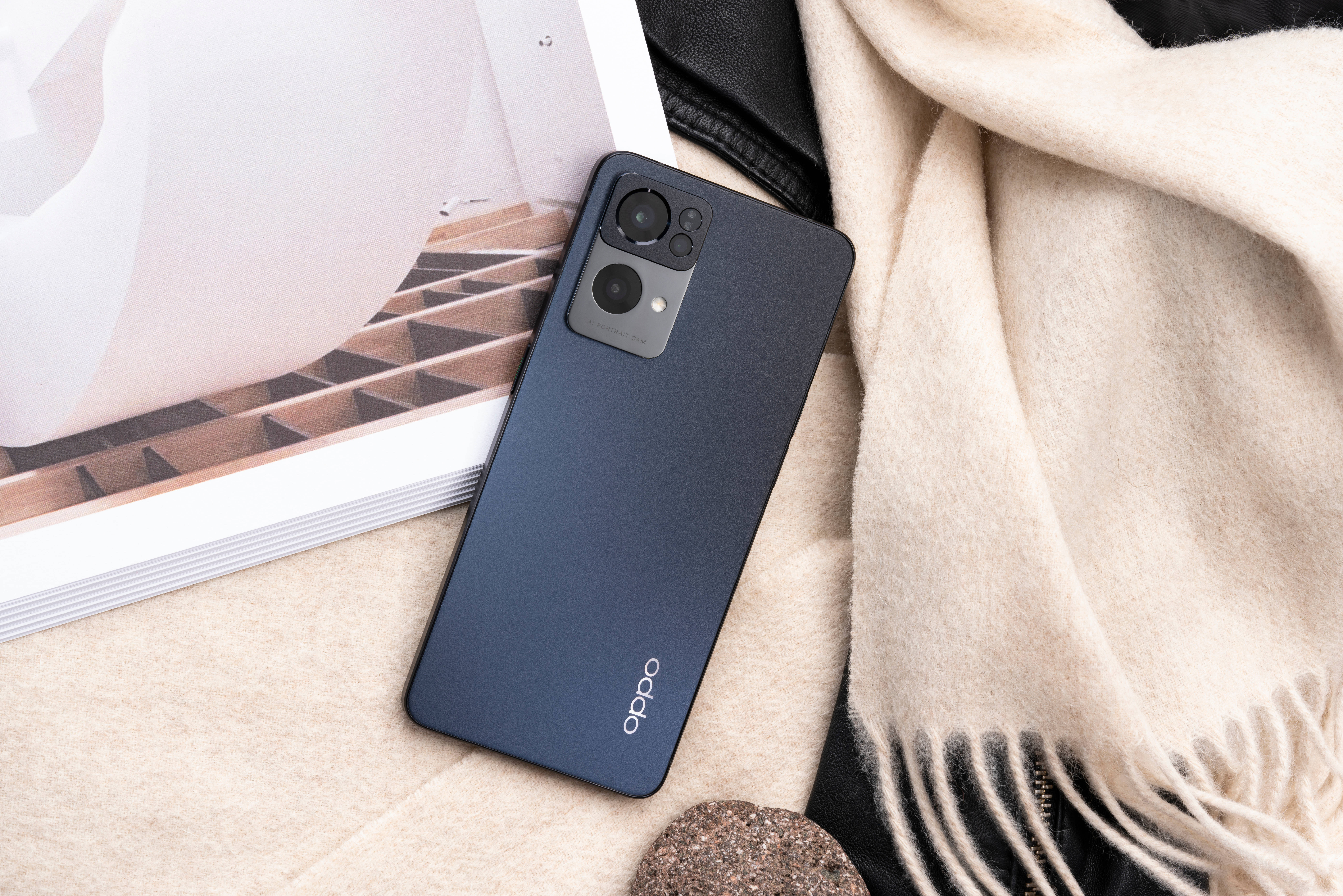 Oppo 桌布 壁紙 — 一部時尚的深藍色 OPPO 智慧型手機放在柔軟的米色毛毯上，旁邊有一本雜誌和帶紋理的配件。