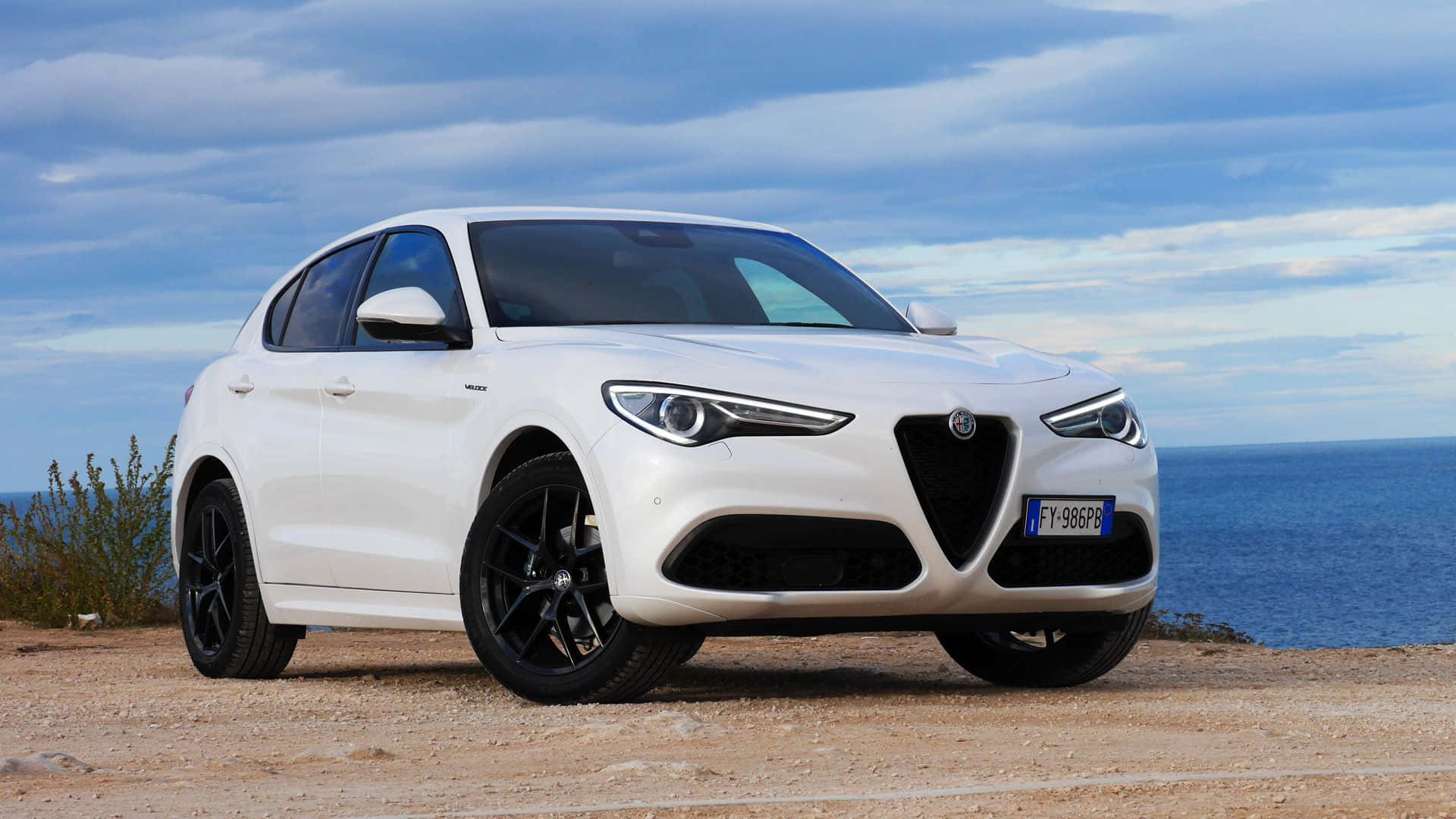Alfa Romeo Stelvio 桌布 壁紙 — 一部時尚的白色 Alfa Romeo SUV 停泊在海岸懸崖上，俯瞰著藍色大海，天空部分多雲。