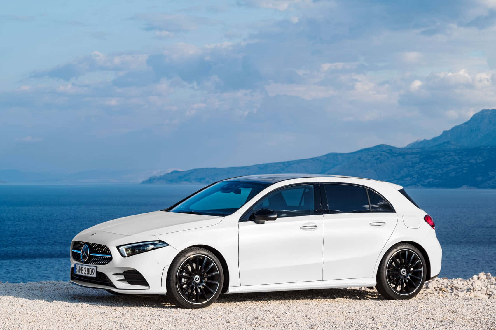 Mercedes Benz A-class 桌布 壁紙 — 一部時尚的白色賓士A-Class掀背車停泊在卵石灘上，背景是廣闊的藍色大海和遠山，天空部分多雲。