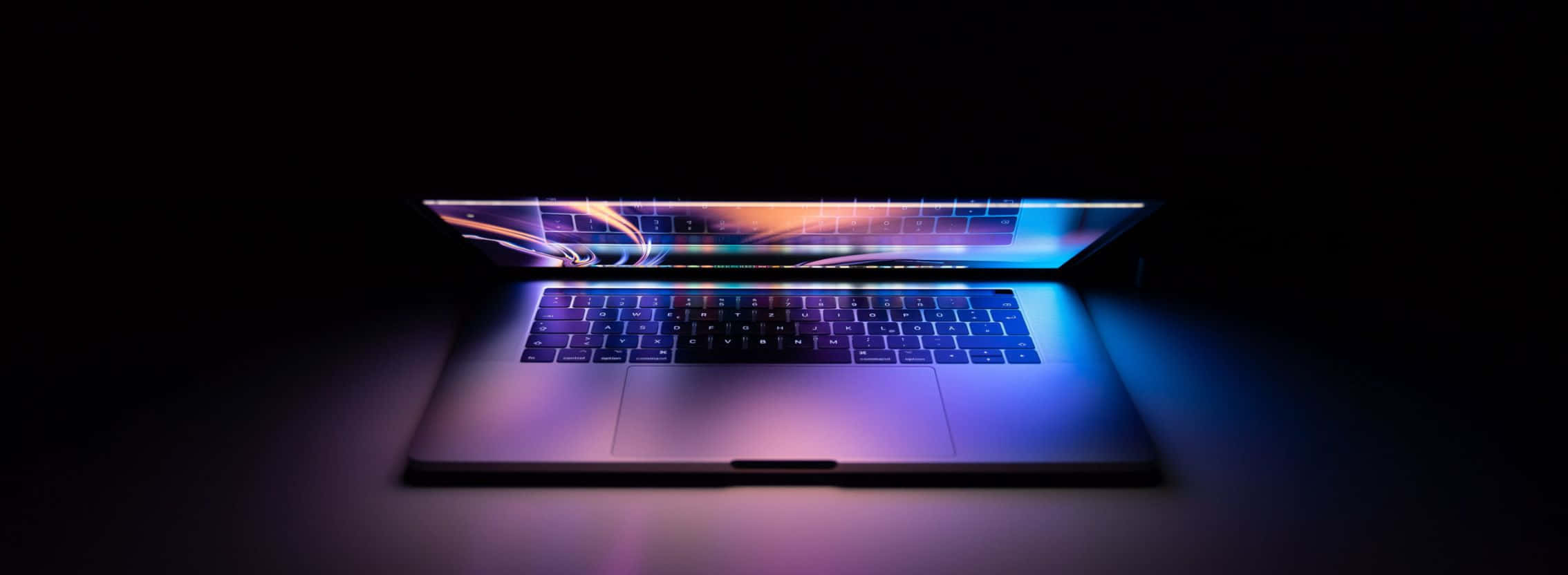 2268x832 桌布 壁紙 — 一部時尚的筆記型電腦在黑暗背景下散發出鮮豔的藍色和紫色光芒。