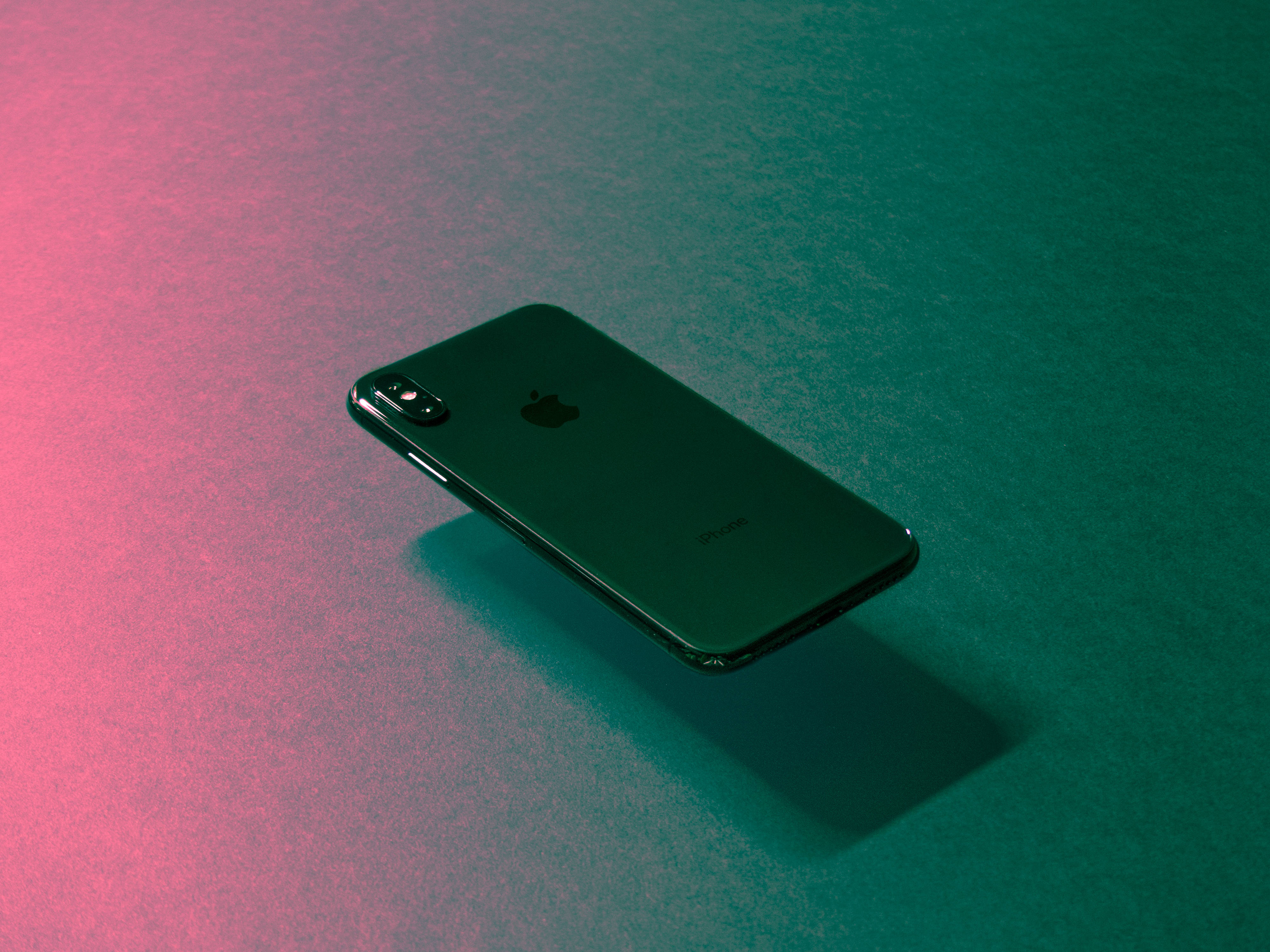 black apple iPhone 桌布 壁紙 — 一部時尚的綠色iPhone對角放置在紋理表面上，沐浴在充滿活力的粉紅色和藍綠色漸變燈光中。