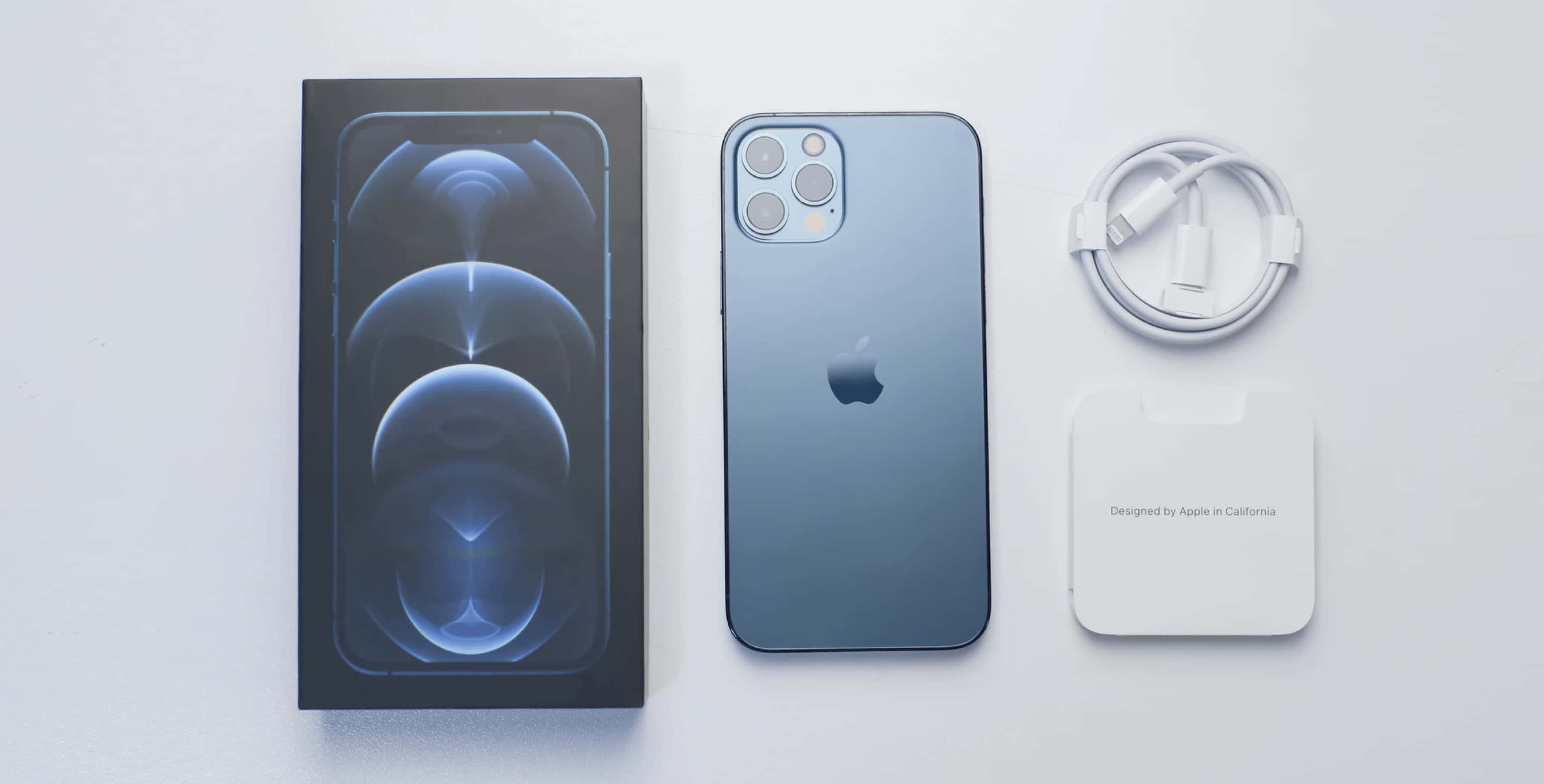 Apple Phone 桌布 壁紙 — 一部時尚的藍色 iPhone 12 Pro 連同其盒裝充電器和線纜放置在簡約的白色表面上。