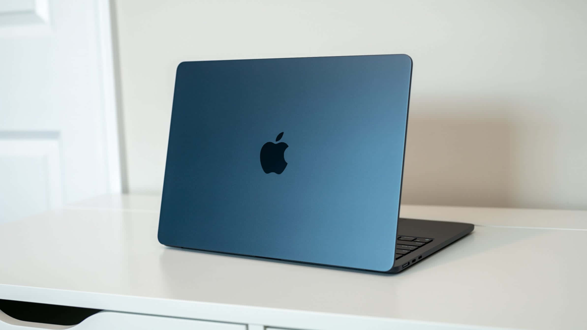 MacBook 動態圖片 桌布 壁紙 — 一部時尚的藍色 MacBook 打開放在白色桌子上，柔和自然光突顯其啞光質感。