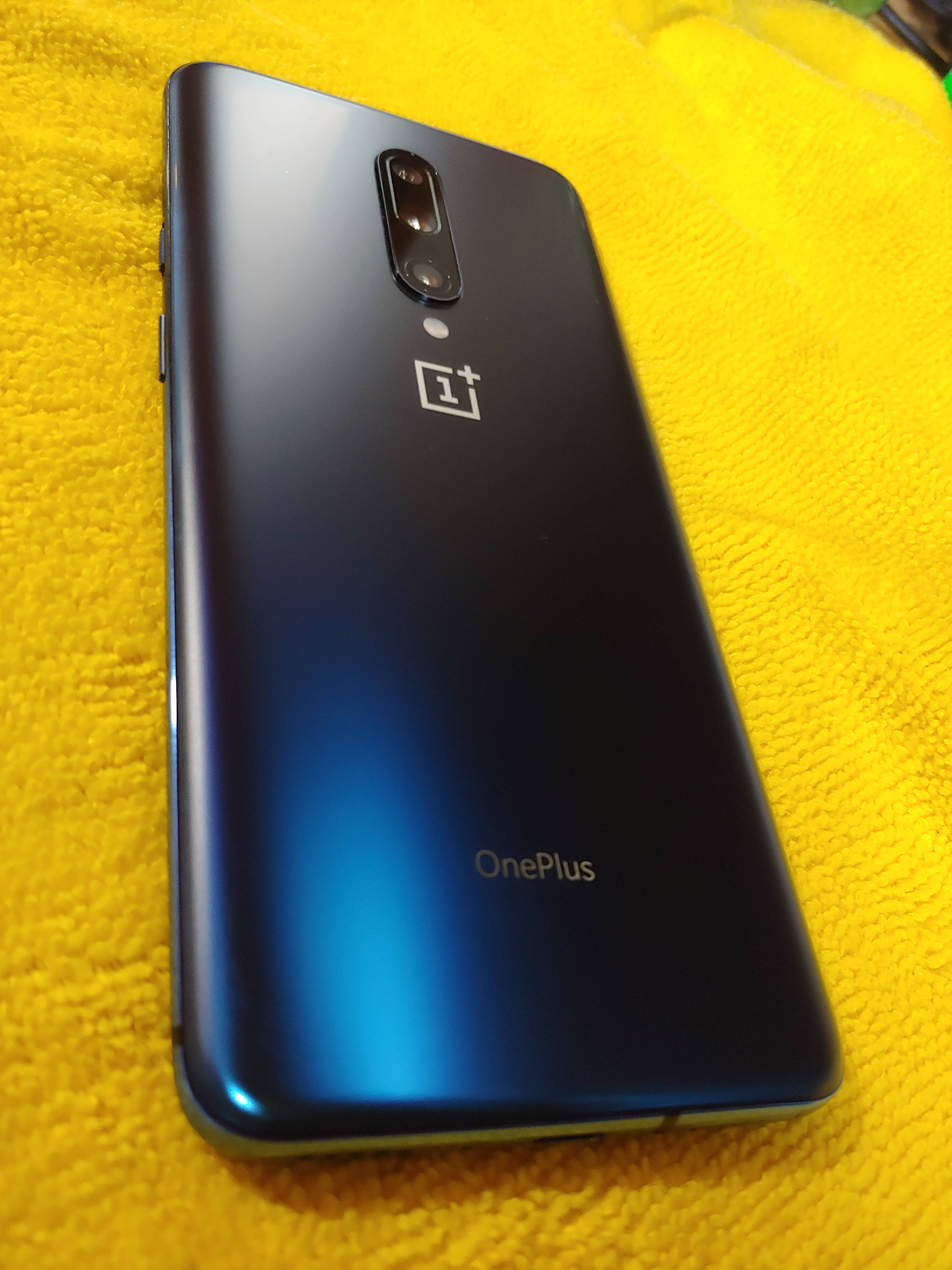  壁紙 — 一部時尚的藍色 OnePlus 智慧型手機放在鮮黃色紋理毛巾上，相機和標誌清晰可見。