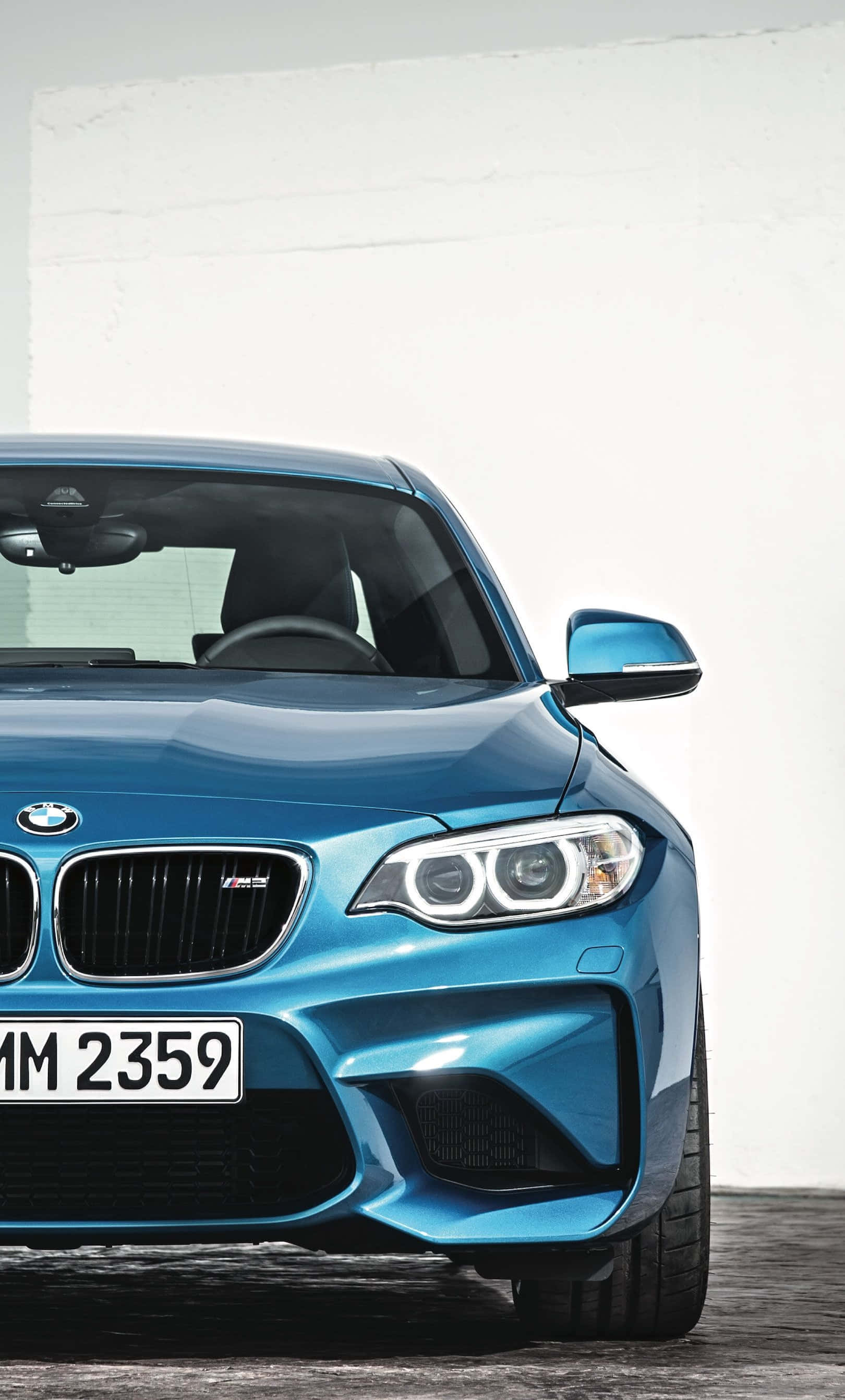 BMW 動圖 桌布 壁紙 — 一部時尚的藍色BMW M2跑車，具有侵略性的前臉設計和發光的頭燈，在簡約的白色牆壁前蓄勢待發。