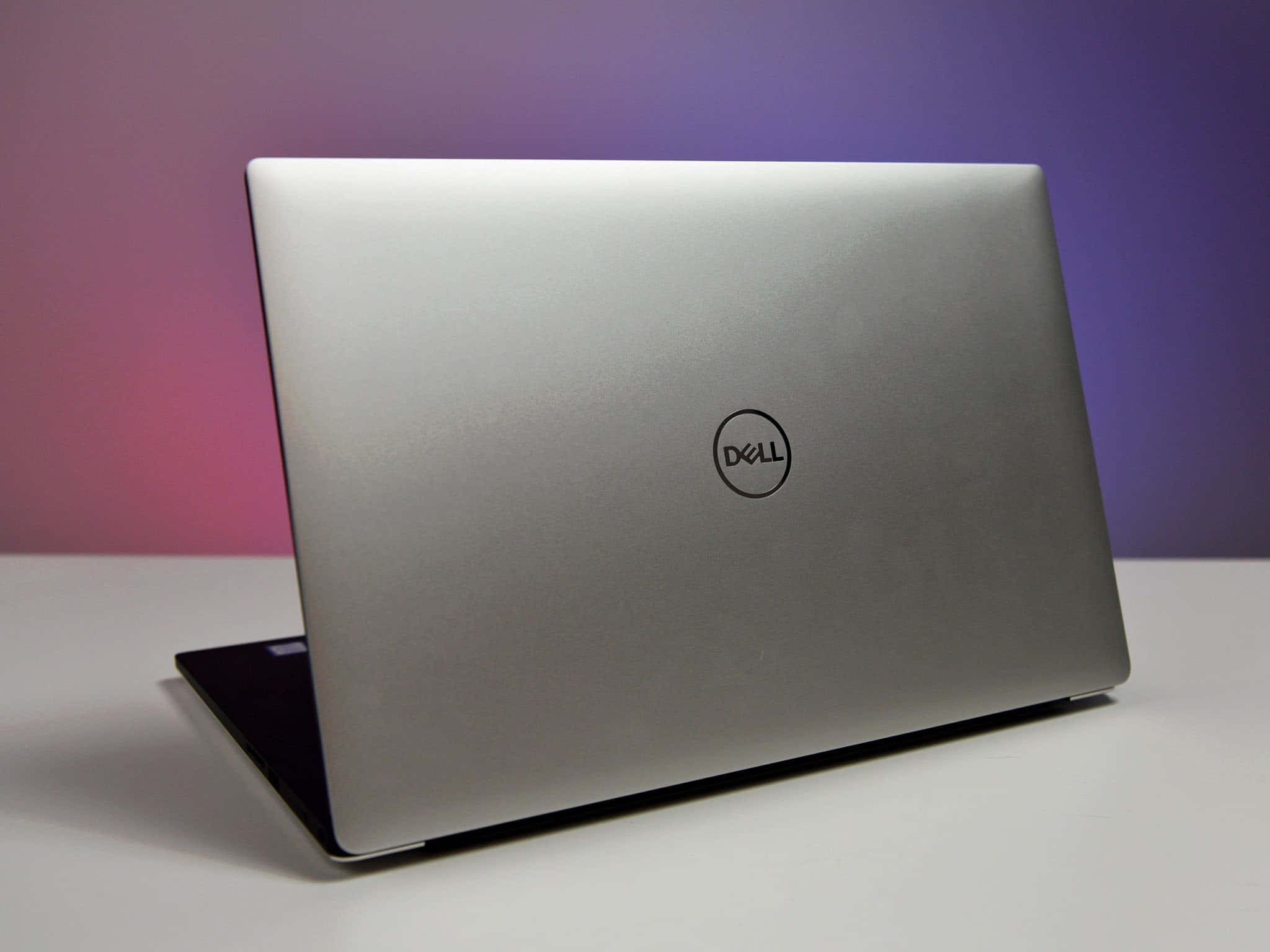 Dell 桌布 壁紙 — 一部時尚的銀色 Dell 筆記型電腦放在白色表面上，背景是粉紅和紫色的漸變色。