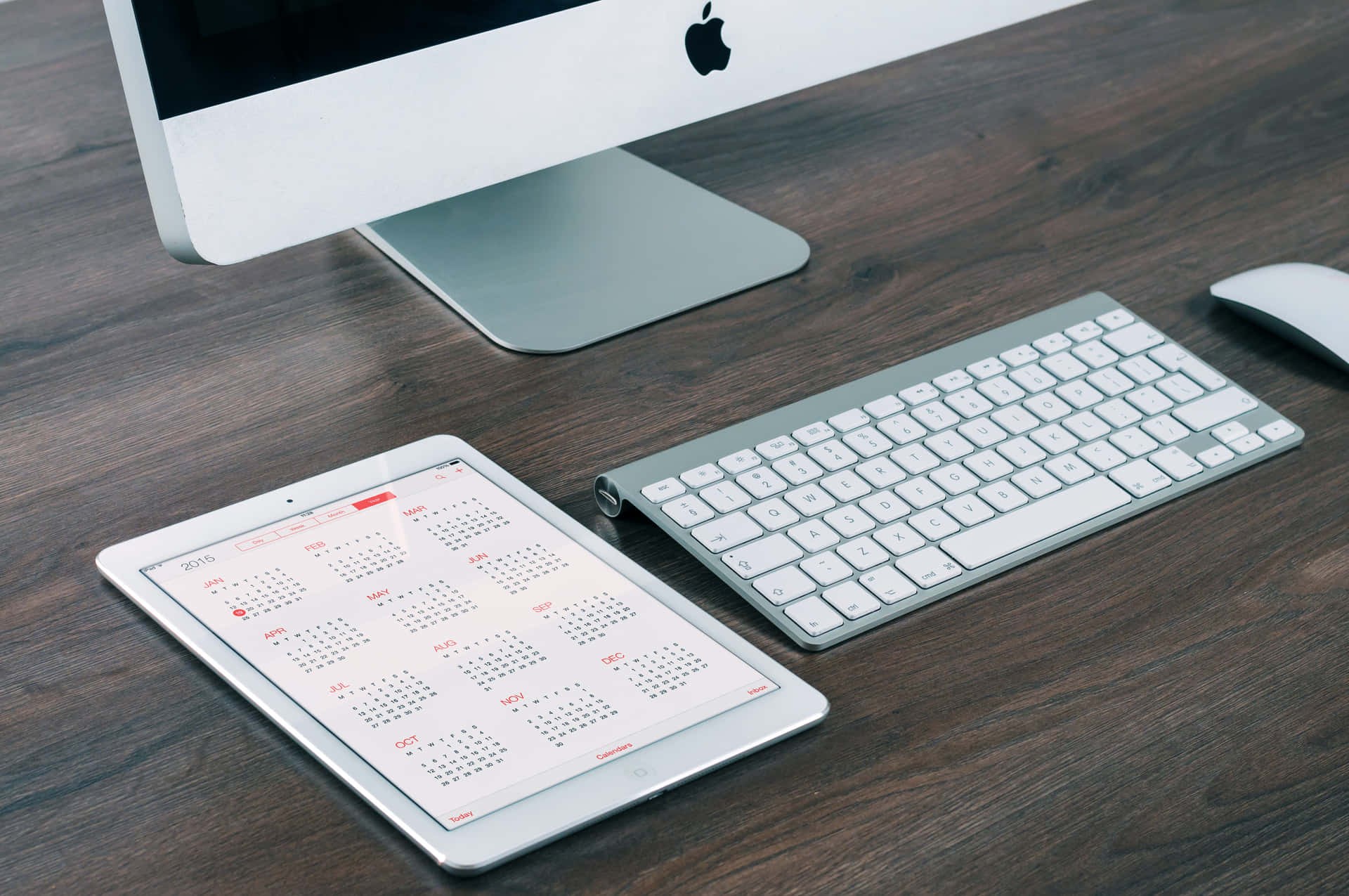 Computer Monitor 桌布 壁紙 — 一部時尚的銀色 iMac、iMac 鍵盤和顯示日曆的 iPad 放在深色木桌上。