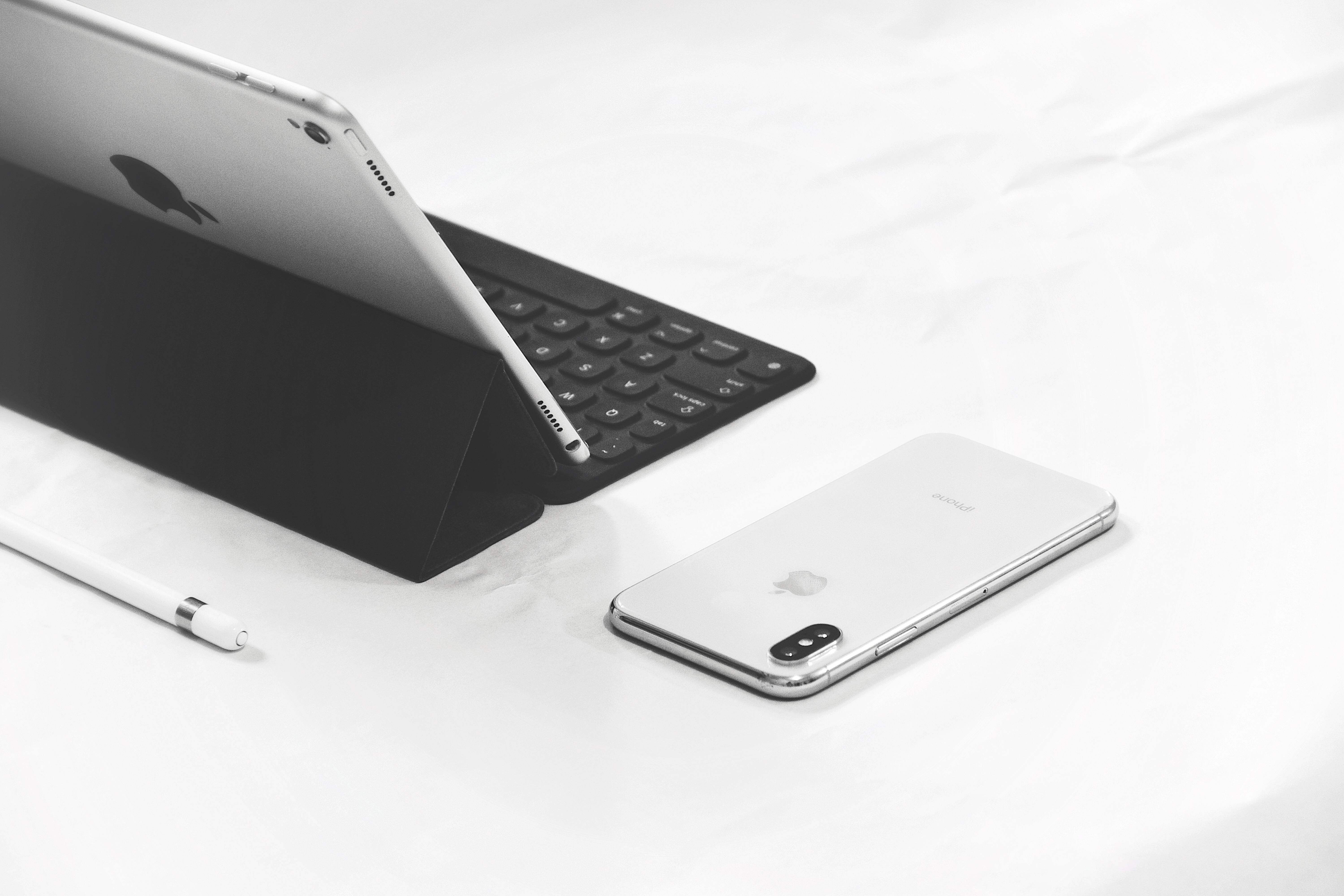 Apple iPad Pro 桌布 壁紙 — 一部時尚的銀色 iPad 配備鍵盤保護套和白色 iPhone 放置在簡約的白色表面上，附近有一支 Apple Pencil。
