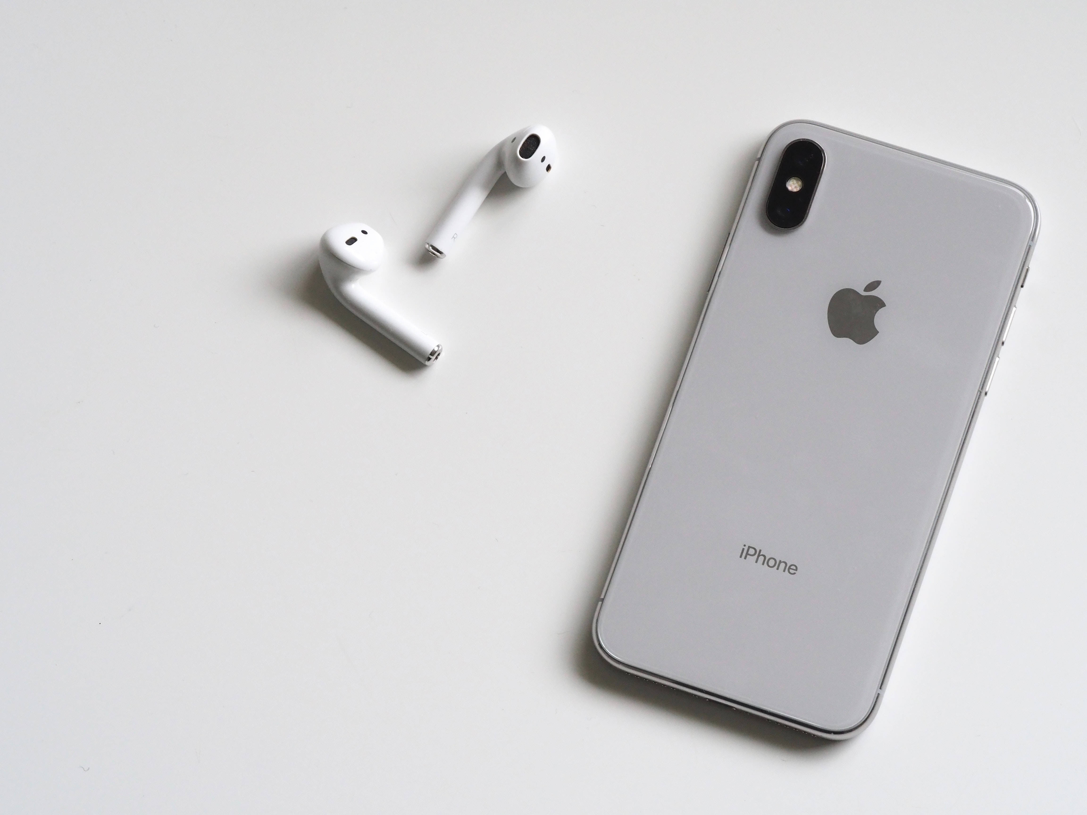 酷炫iPhone 桌布 壁紙 — 一部時尚的銀色 iPhone X 和白色 AirPods 在柔和燈光下放置在簡約的白色表面上。