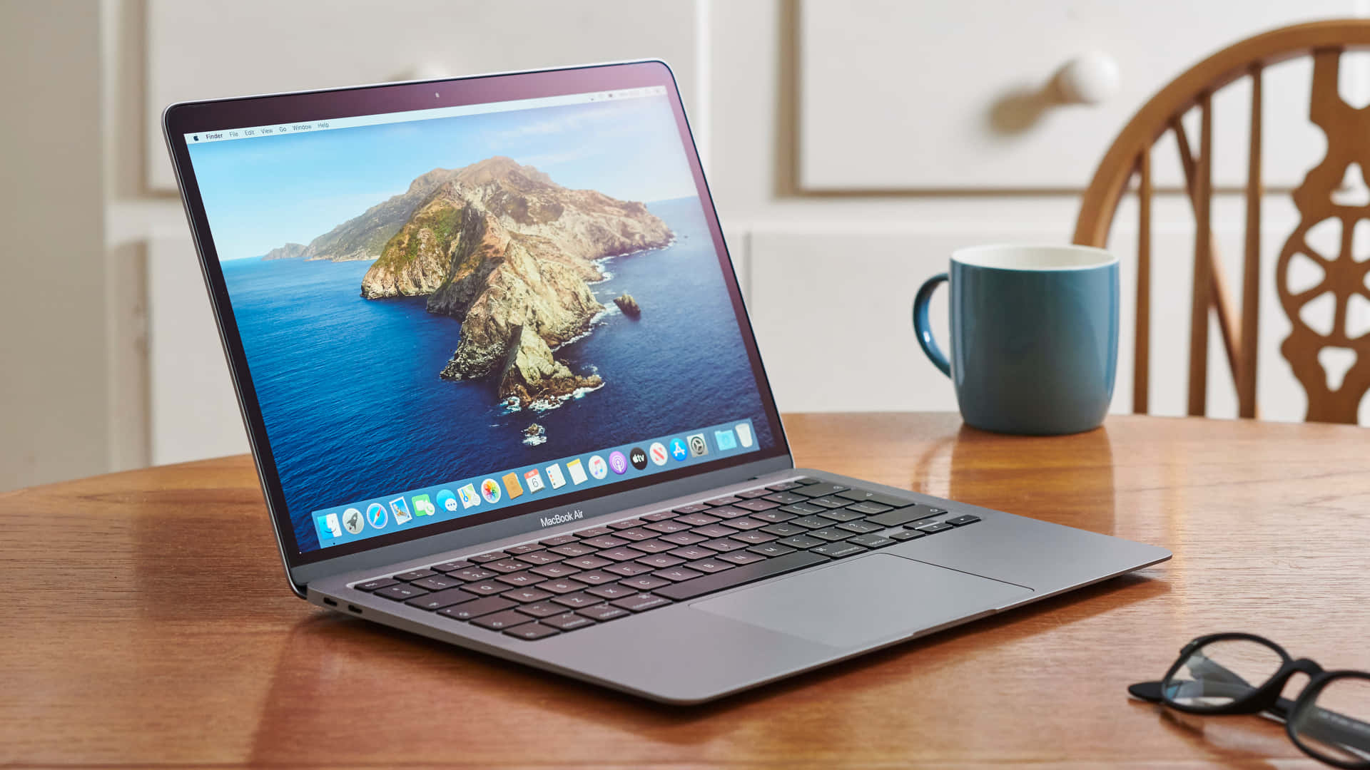 MacBook 動態圖片 桌布 壁紙 — 一部時尚的銀色 MacBook Air 打開放在木桌上，顯示充滿活力的海岸島嶼壁紙，旁邊有一個藍色杯子和眼鏡。