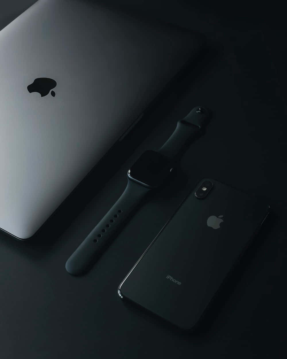 iPhone 桌布 壁紙 — 一部時尚的銀色 MacBook 筆記型電腦、黑色 Apple Watch 和黑色 iPhone 在柔和燈光下排列在深色表面上。