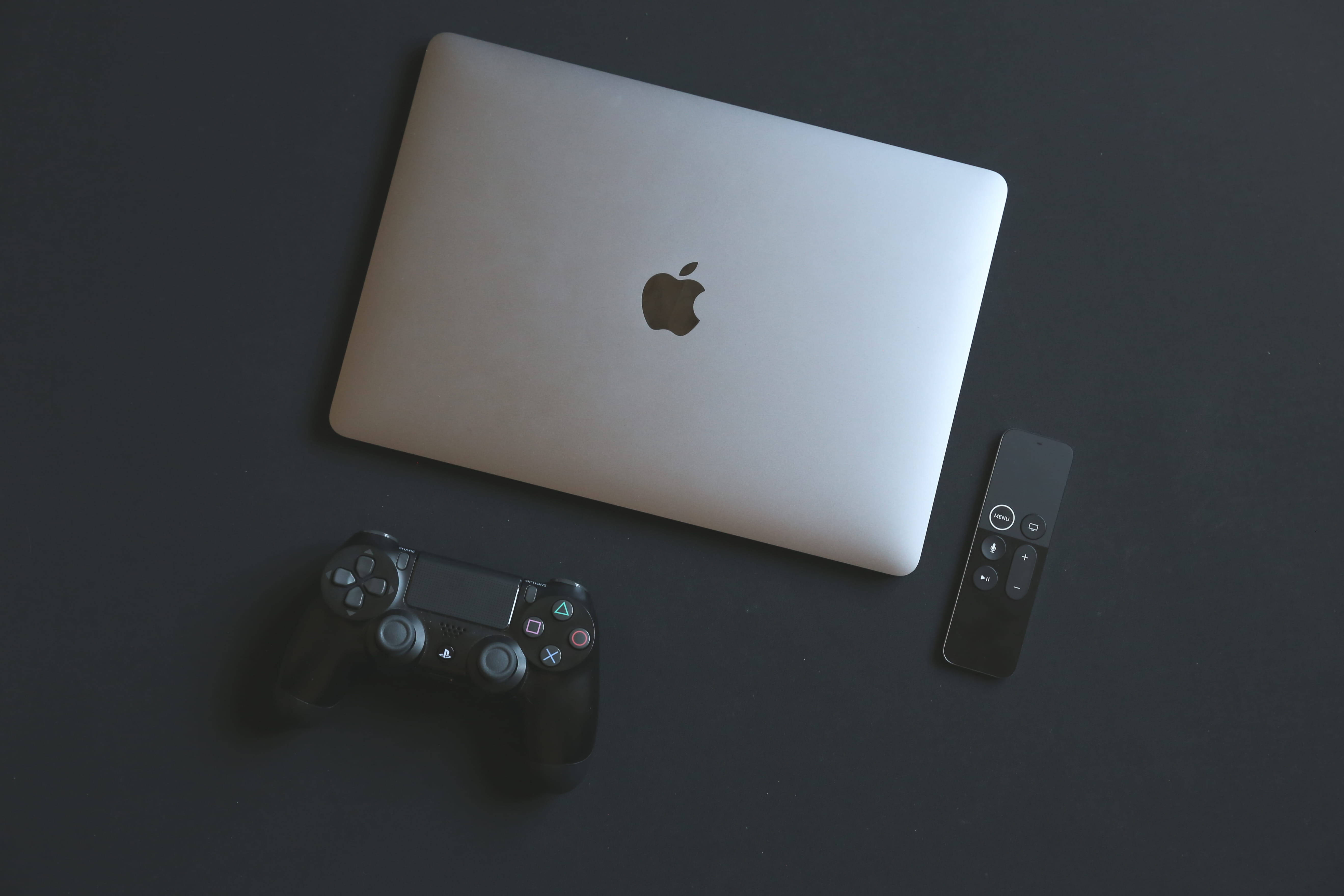 PlayStation 桌布 壁紙 — 一部時尚的銀色 MacBook 筆記型電腦旁邊放著一個黑色 PlayStation 控制器和 Apple TV 遙控器，置於深色表面上。