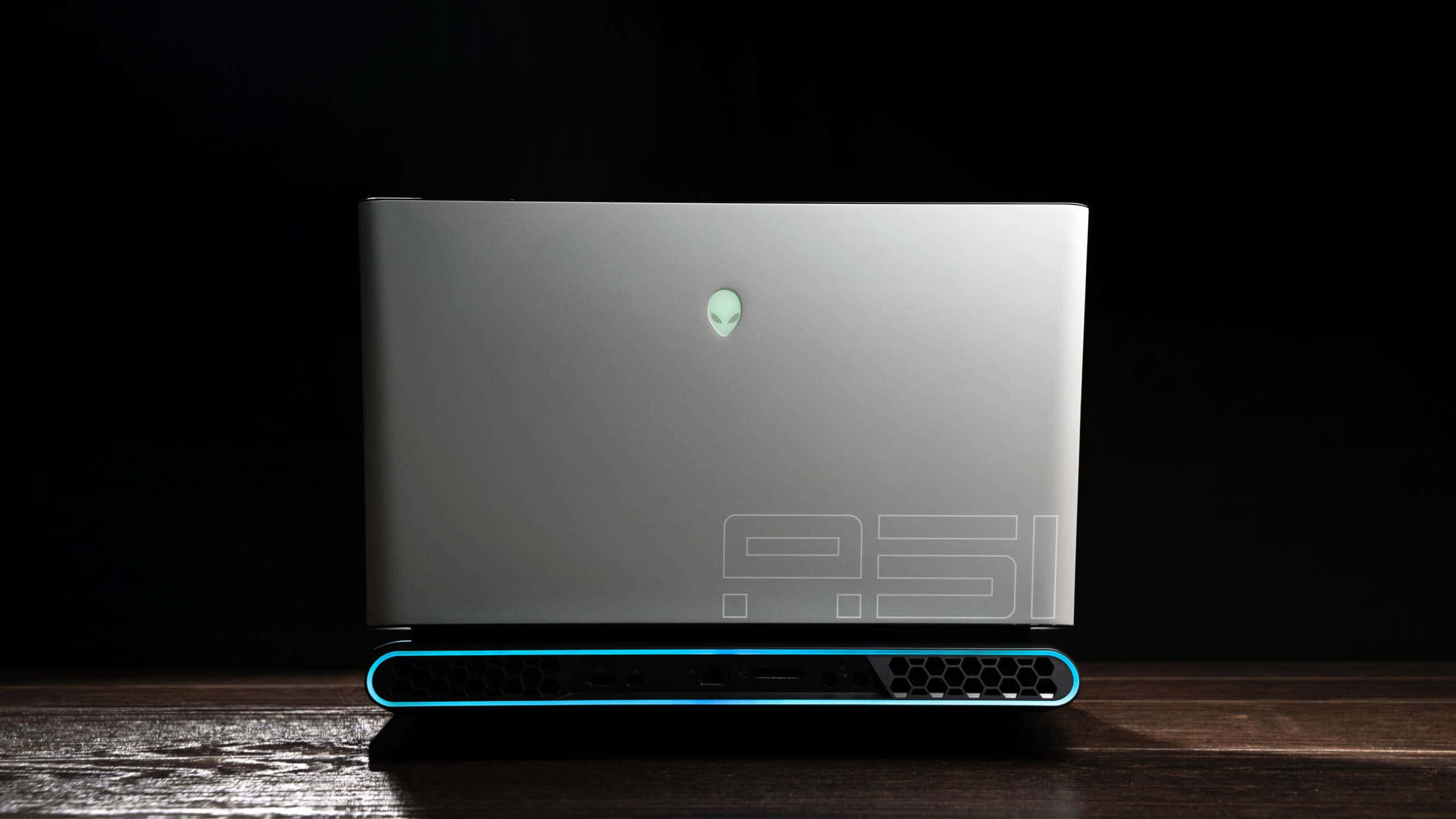 外星人 桌布 壁紙 — 一部時尚的銀色Alienware筆記型電腦，帶有發光的藍色點綴，關閉在黑色背景下的深色木質表面上。
